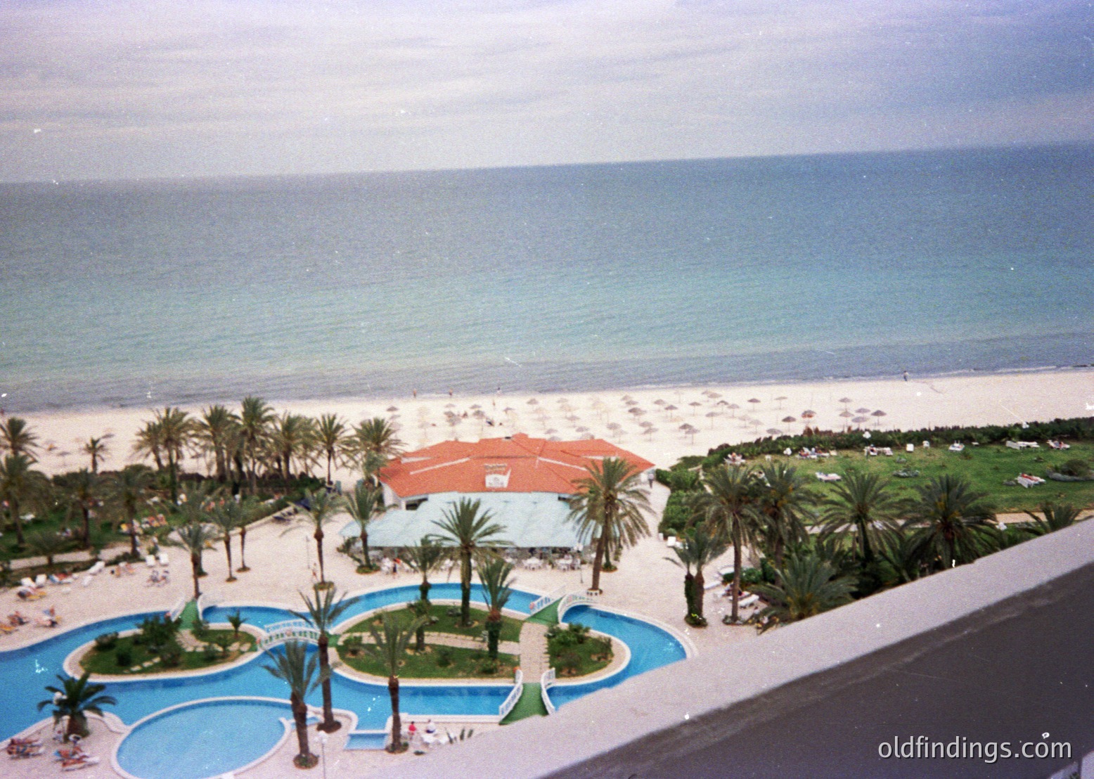 Mid-20th century seaside resort with tiered pools, palm-dotted gardens, and oceanfront beach. Architectural style suggests 1960s-70s Mediterranean or tropical resort design.
