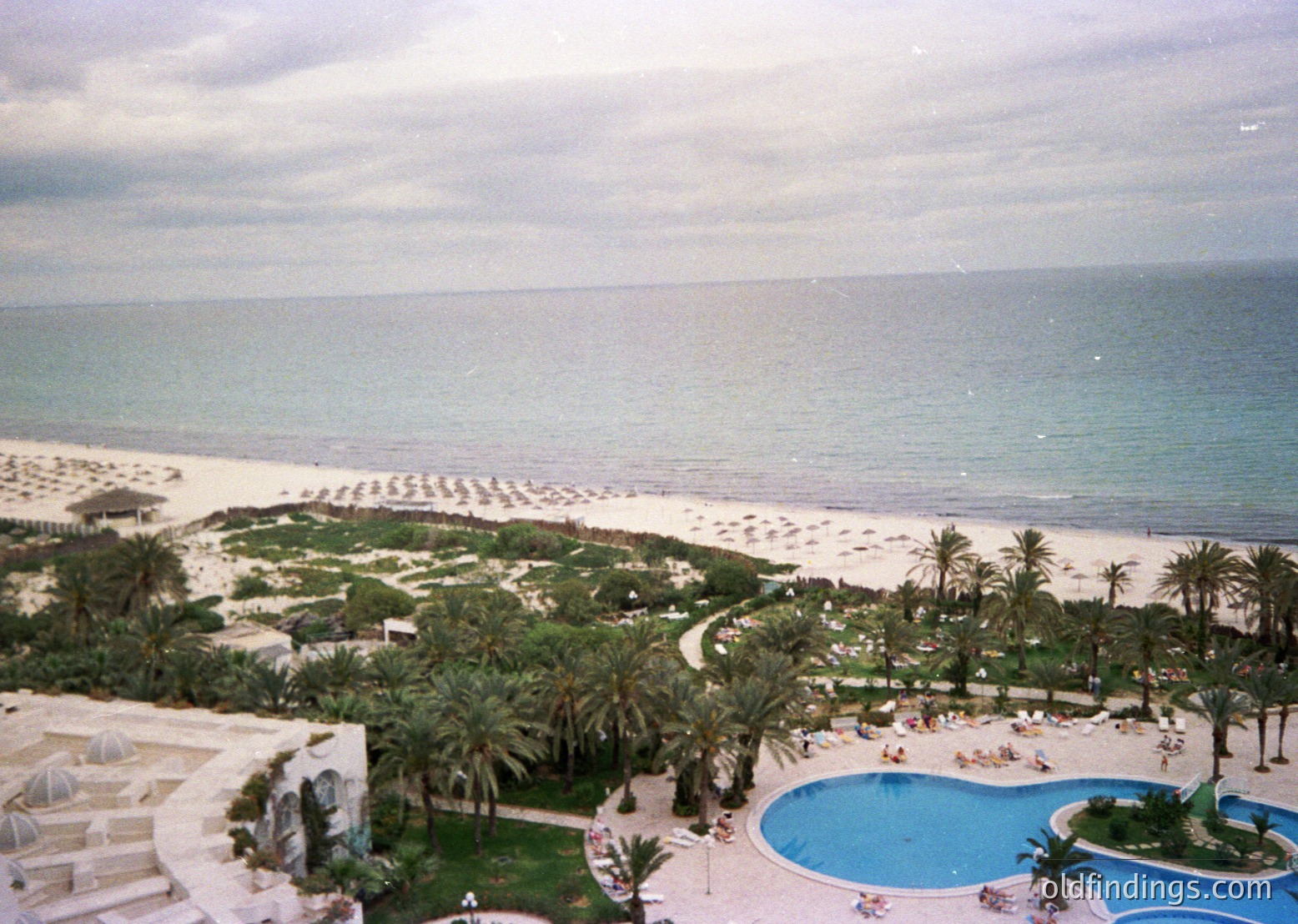 Mid-20th-century seaside resort with curved infinity pool overlooking golden beach and turquoise waters. Palm-dotted lawns, sun loungers, and shaded umbrellas frame the scene. Architectural style suggests Mediterranean influence, likely or .