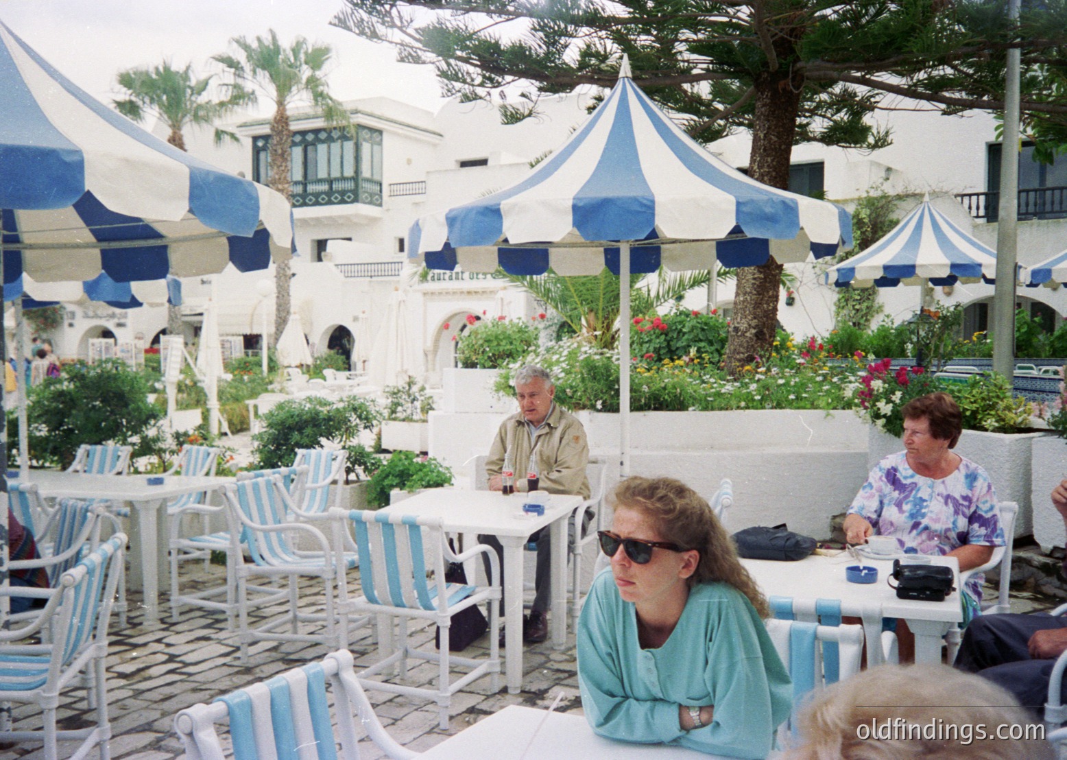 Mid-1990s seaside café with Mediterranean architecture—whitewashed walls, arched doorways, and lush greenery. Patrons dine under striped blue-and-white umbrellas at striped tables/chairs. Casual summer attire (sunglasses, swimwear, light layers) suggests a warm climate. Likely or . é