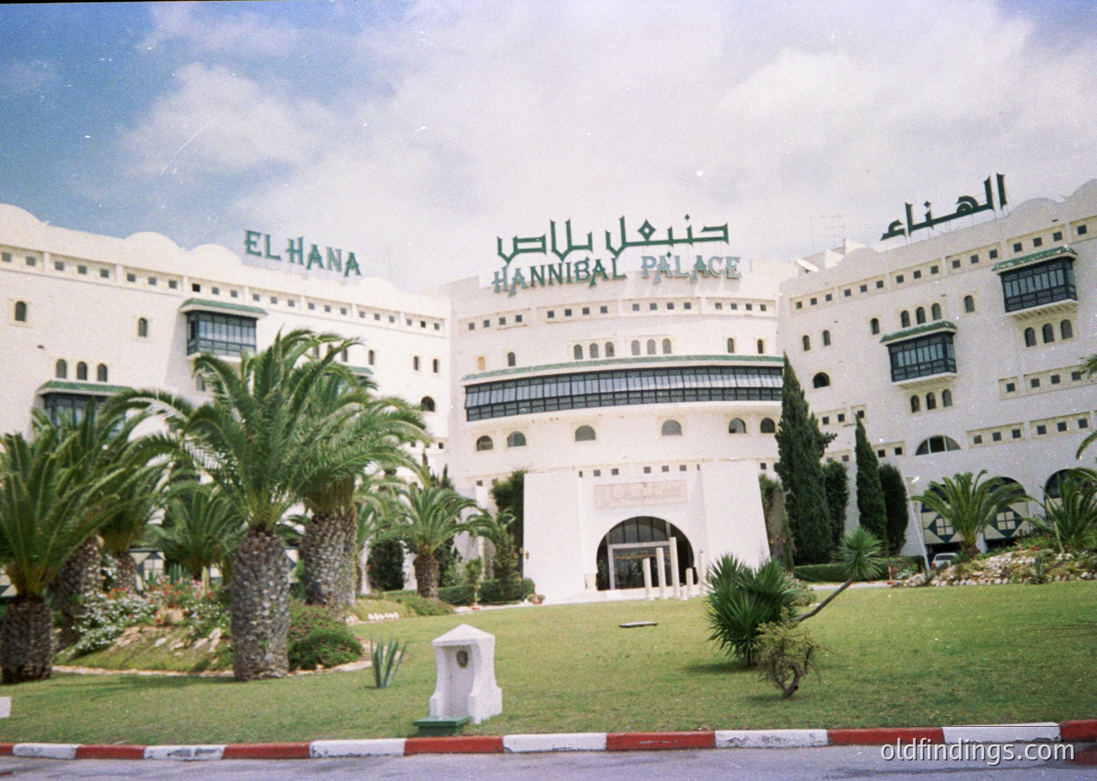 Mid-century luxury hotel facade featuring Arabic script ("El Hana" & "Hannibal Palace") in bold, curved signage. Whitewashed walls with arched windows, balconies, and decorative palm trees in landscaped courtyard. Likely --- *Note: The architectural style and script suggest a location in Jordan, likely Amman, during the mid-20th century.*