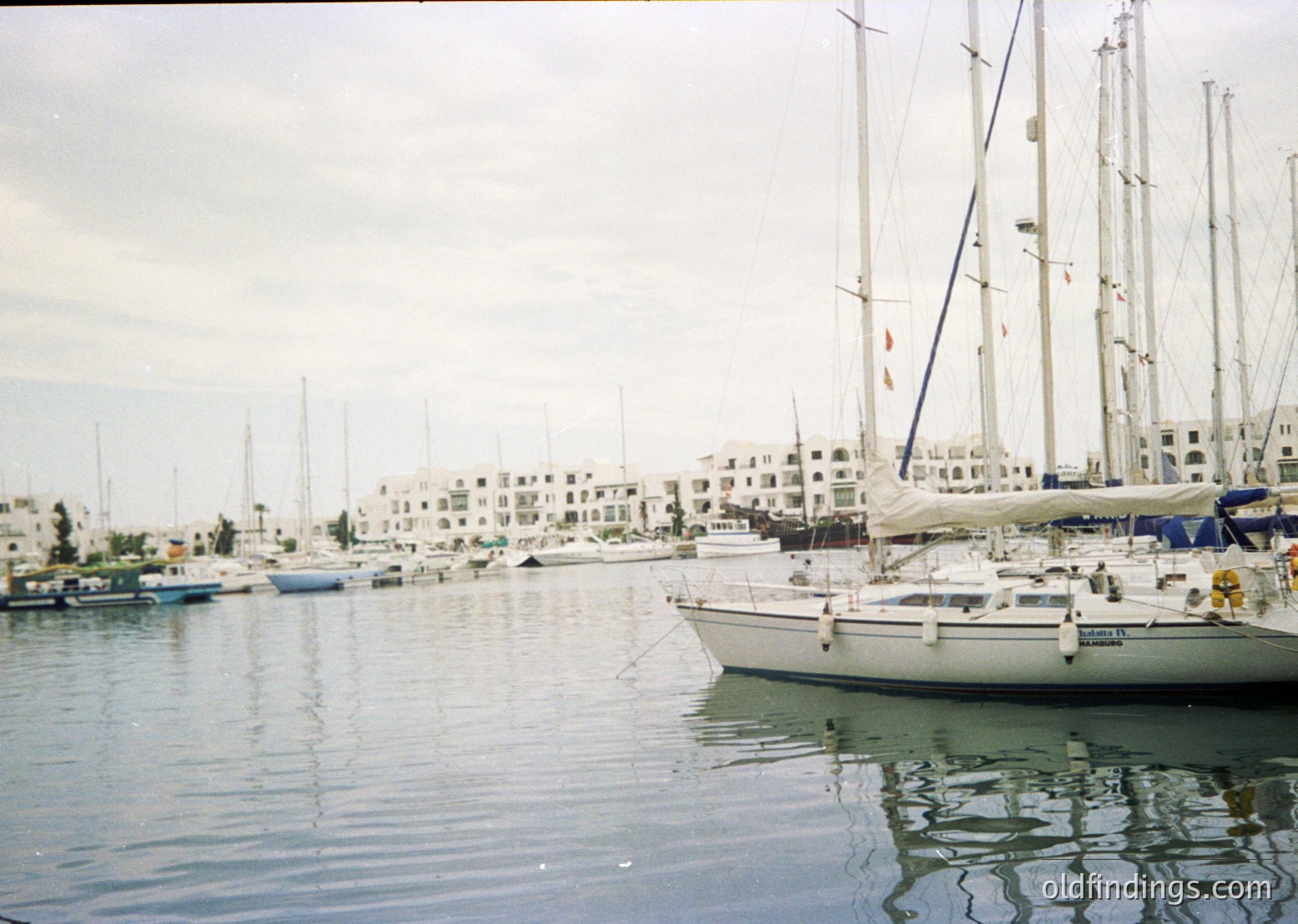 Marina scene with classic sailboats docked alongside Mediterranean-style waterfront buildings. Reflective water and masts dominate foreground. Likely 1990s-2000s Mediterranean coastal town.