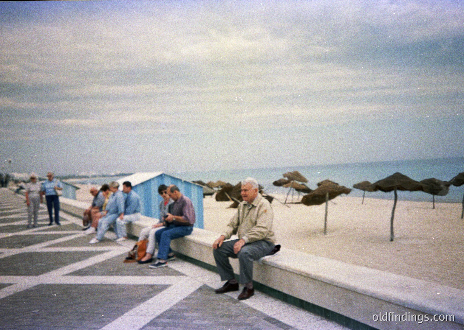 Vintage seaside promenade with mid-century concrete seating. Elderly man in light jacket sits on a curved concrete ledge, overlooking the Mediterranean. Blue beach huts and straw umbrellas line the shore. Overcast sky suggests early 1970s European coastal vibe.