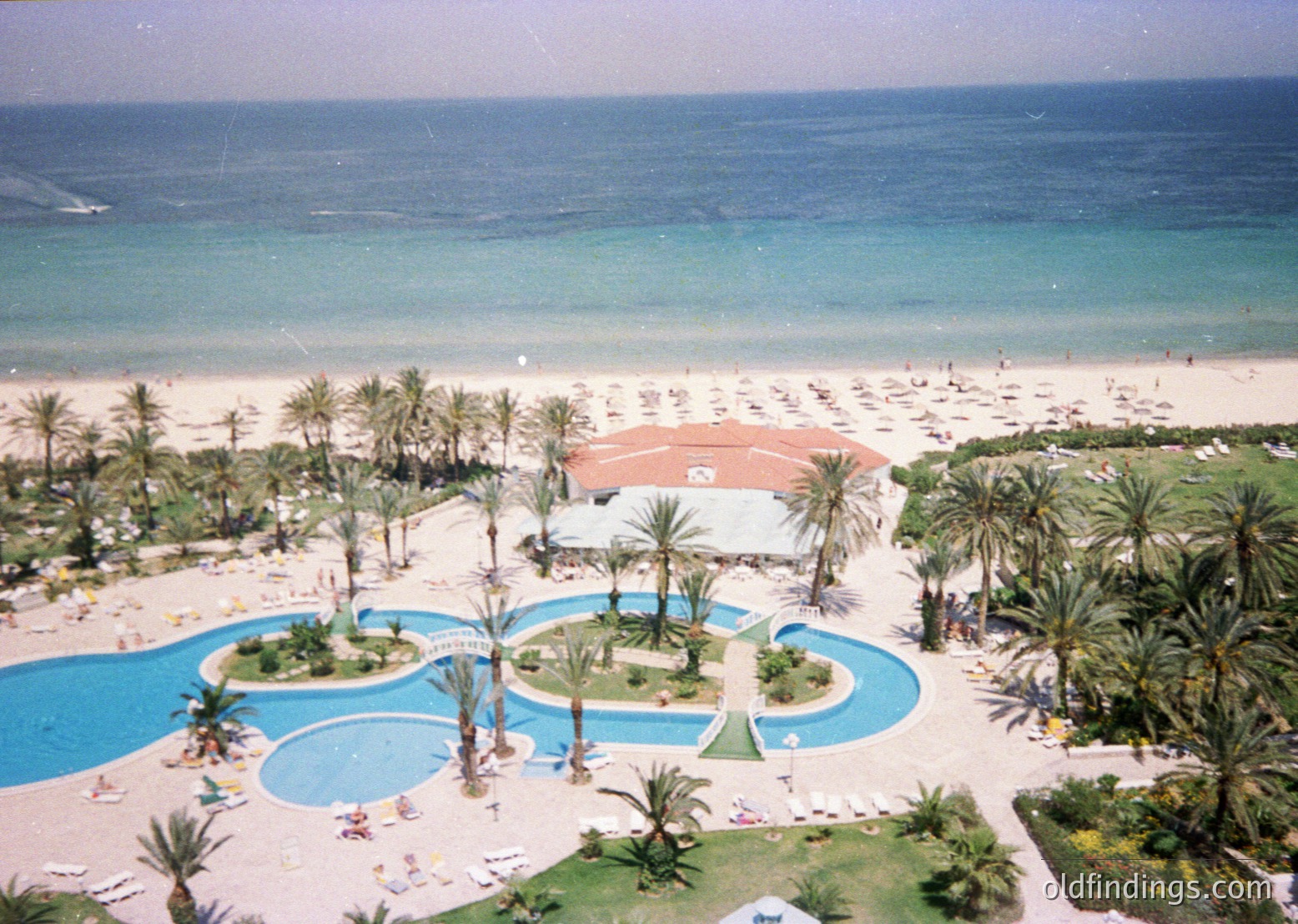 Aerial view of a mid-20th-century seaside resort with tiered pools, lush palm groves, and turquoise waters. Sunbathers and beachgoers populate the sandy shore. Architectural style blends Mediterranean with mid-century modern.
