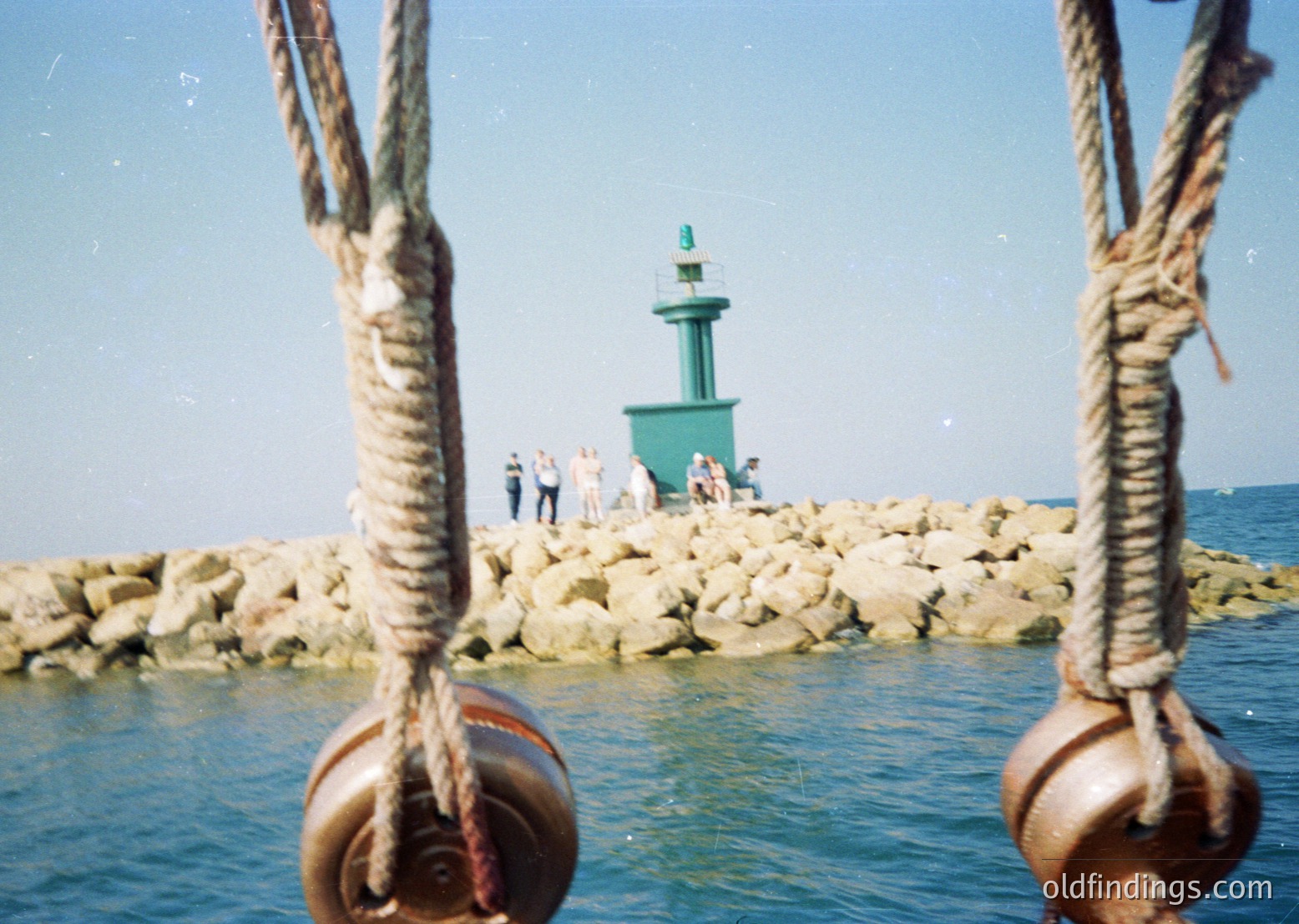 Vintage seaside lighthouse perched on a breakwater of stacked rocks, framed by rusted metal mooring rings. Small group of people in summer attire (1960s-1980s) standing near the base. Clear blue water and sky suggest Mediterranean or coastal European setting.