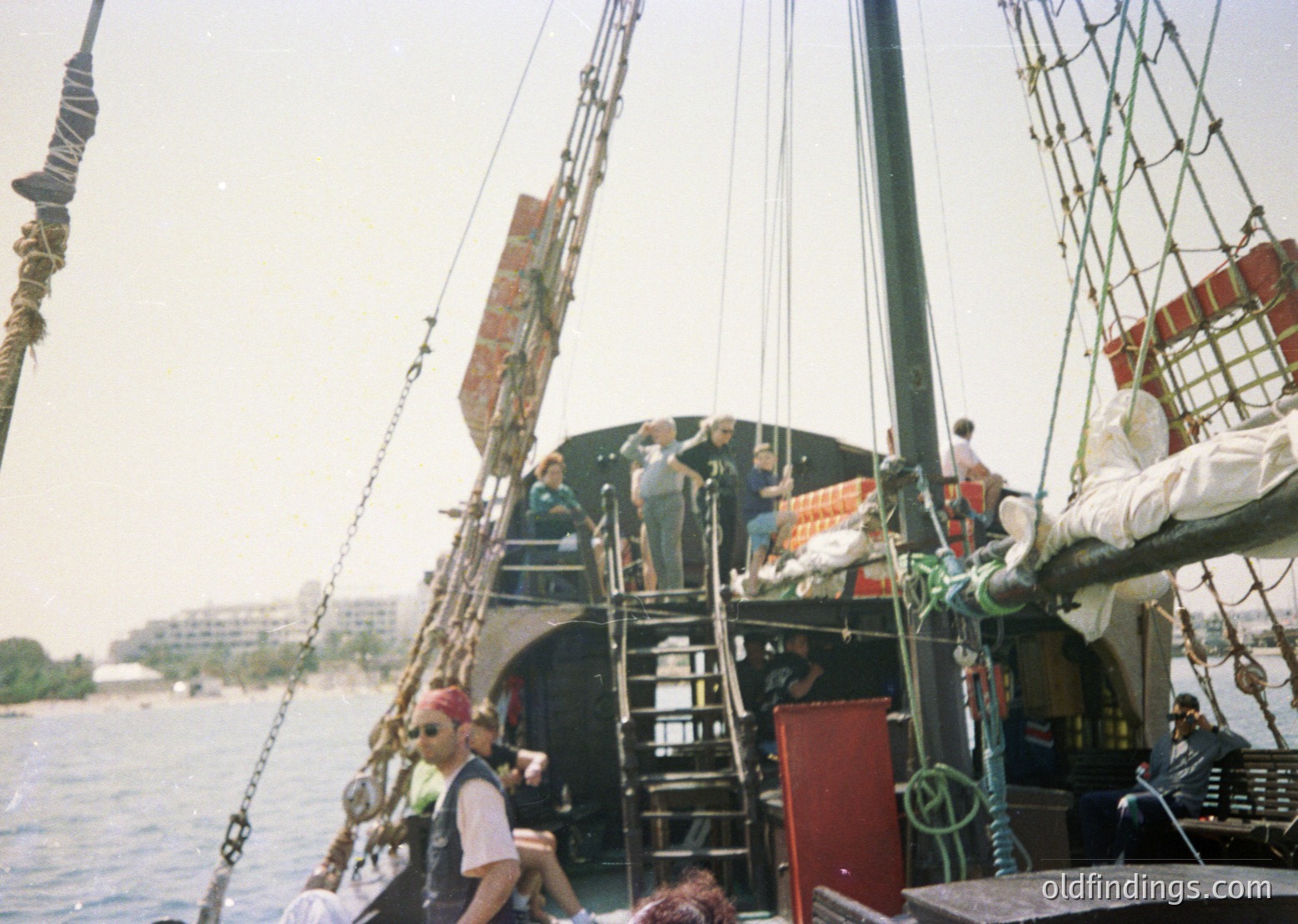 Vintage wooden sailing ship with three masts, likely a replica of a 17th-century East Indiaman, docked near an urban waterfront. Crew in period attire (long sleeves, hats) and modern casual wear (shorts, caps) navigate rigging. Distinctive red-painted cabin door and stacked crates visible. --- *Note: The approximate time period is inferred from the clothing mix and ship style, suggesting a 1970s reenactment or educational voyage.*