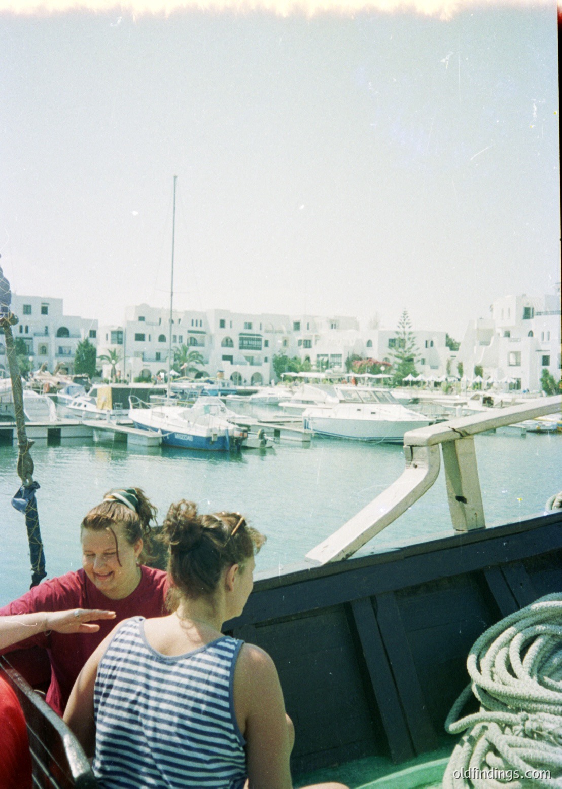 Two women in 1960s-style swimwear stand on a wooden boat docked near a marina with whitewashed Mediterranean-style buildings. The woman in red poses while the striped swimsuit-wearing woman gestures. Yachts and palm trees frame the coastal backdrop.