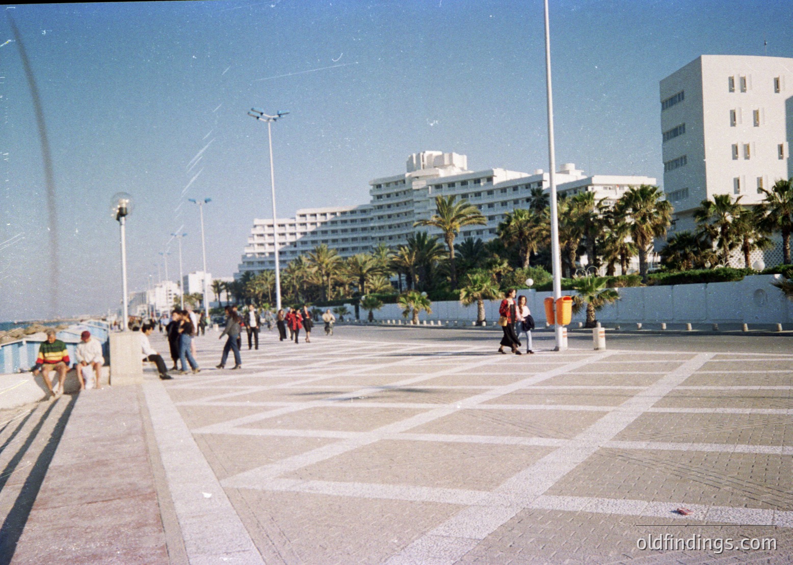 Seaside promenade with geometric patterned pavement, lined by palm trees and mid-century modern hotels. Crowds stroll along the wide walkway under clear skies, with a curved fountain arching over the left side. Likely Mediterranean coastal city, 1960s-1970s era.