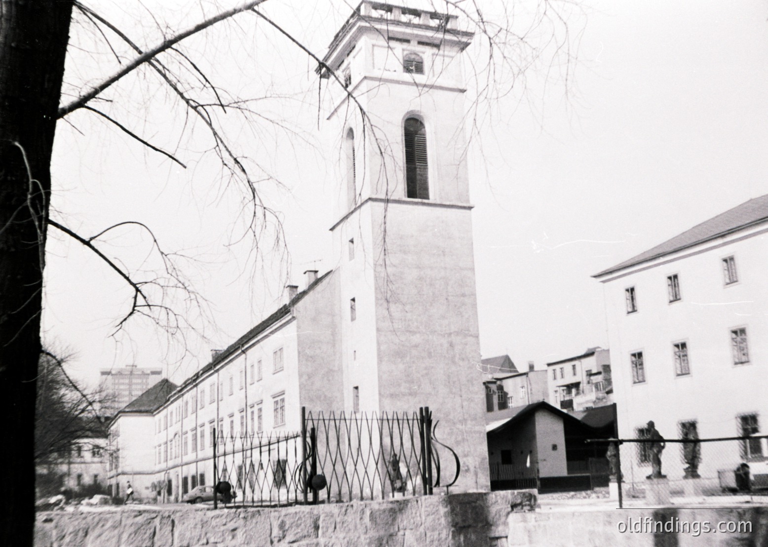 Mid-20th century urban scene featuring a tall, square clock tower with a bell and decorative cornice, likely part of a public or institutional building. Snow covers rooftops and ground, indicating winter. Leafless trees frame the composition. Fenced area with barbed wire suggests restricted access. Architectural style hints at European influence, possibly Eastern Bloc.
