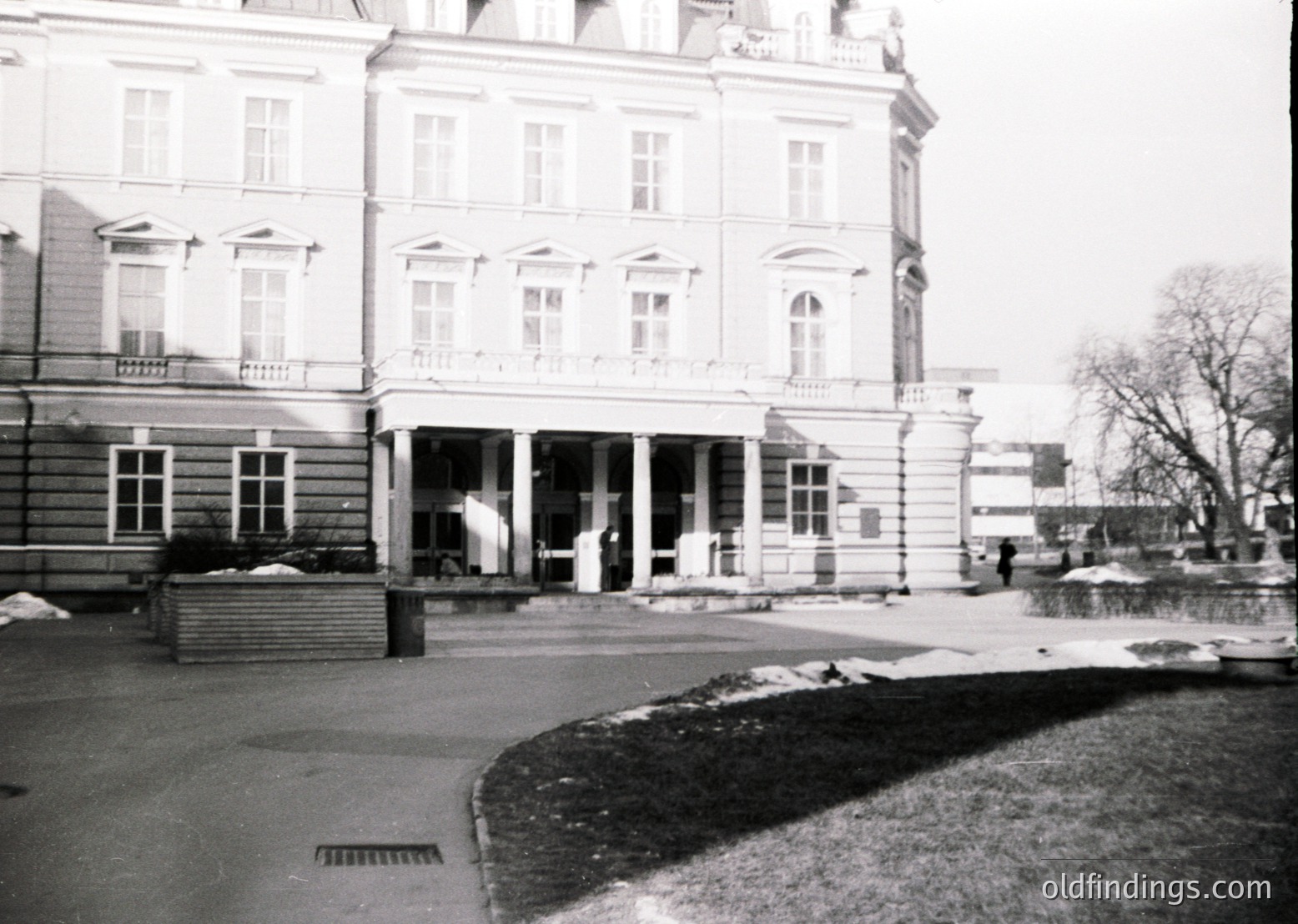 Neoclassical building with symmetrical façade, arched entrance, and balustraded balcony—likely a public or institutional structure. Snow patches and bare trees suggest winter. Mid-20th century European architecture, possibly Eastern Bloc era.