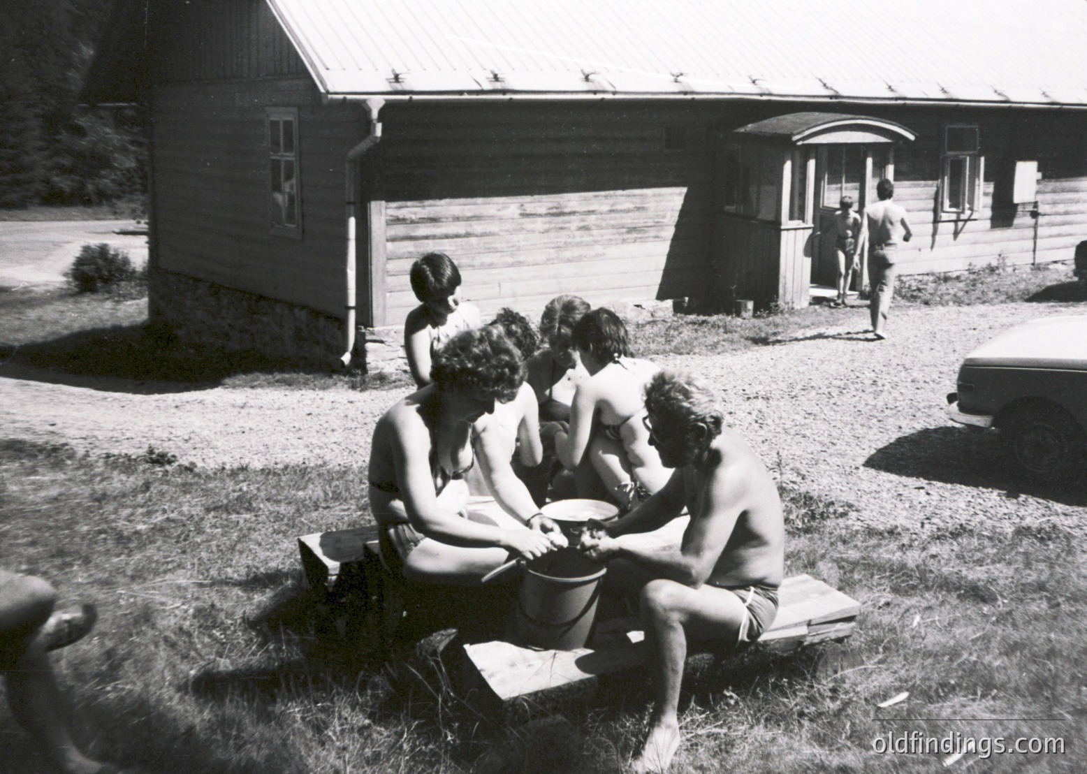 Mid-20th century group photo: five individuals in swimwear playing with a bucket near a rustic wooden cabin. Classic 1950s-60s summer scene with vintage car parked in background.