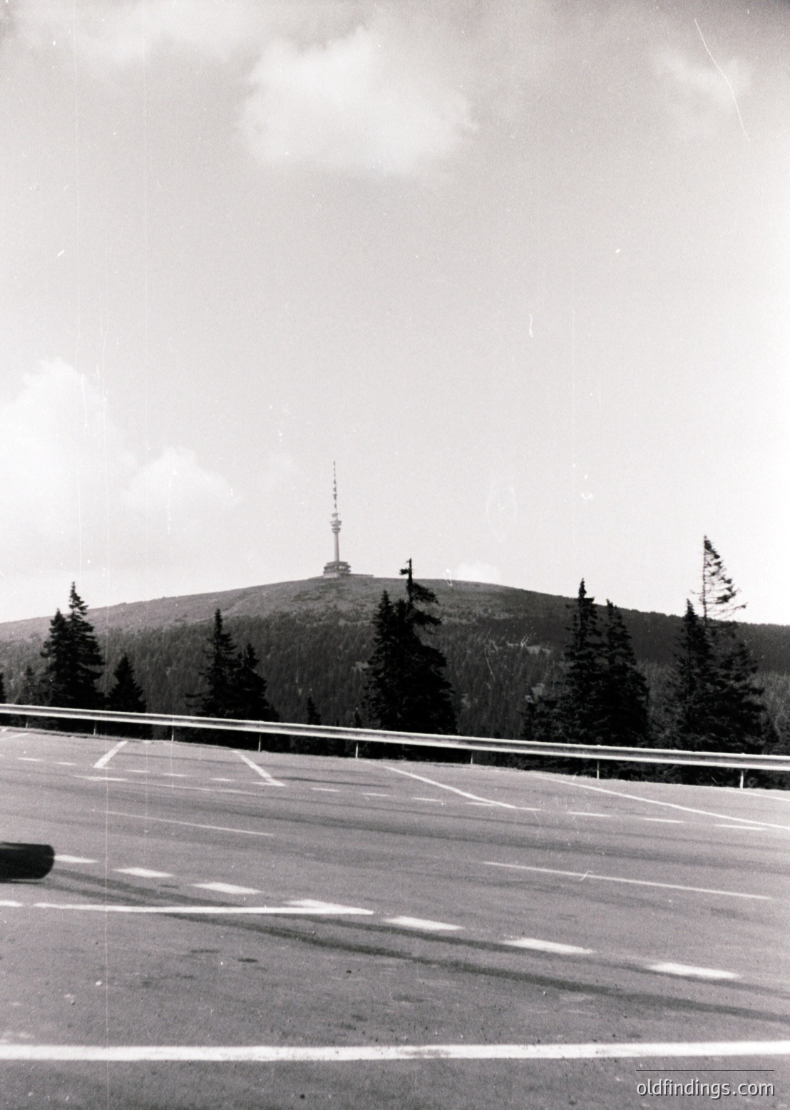 Mid-century roadside view of a tower atop a forested hill, likely a telecommunications or observation structure. Asphalt road with guardrail and parking spaces in foreground. Dense coniferous trees frame the base.