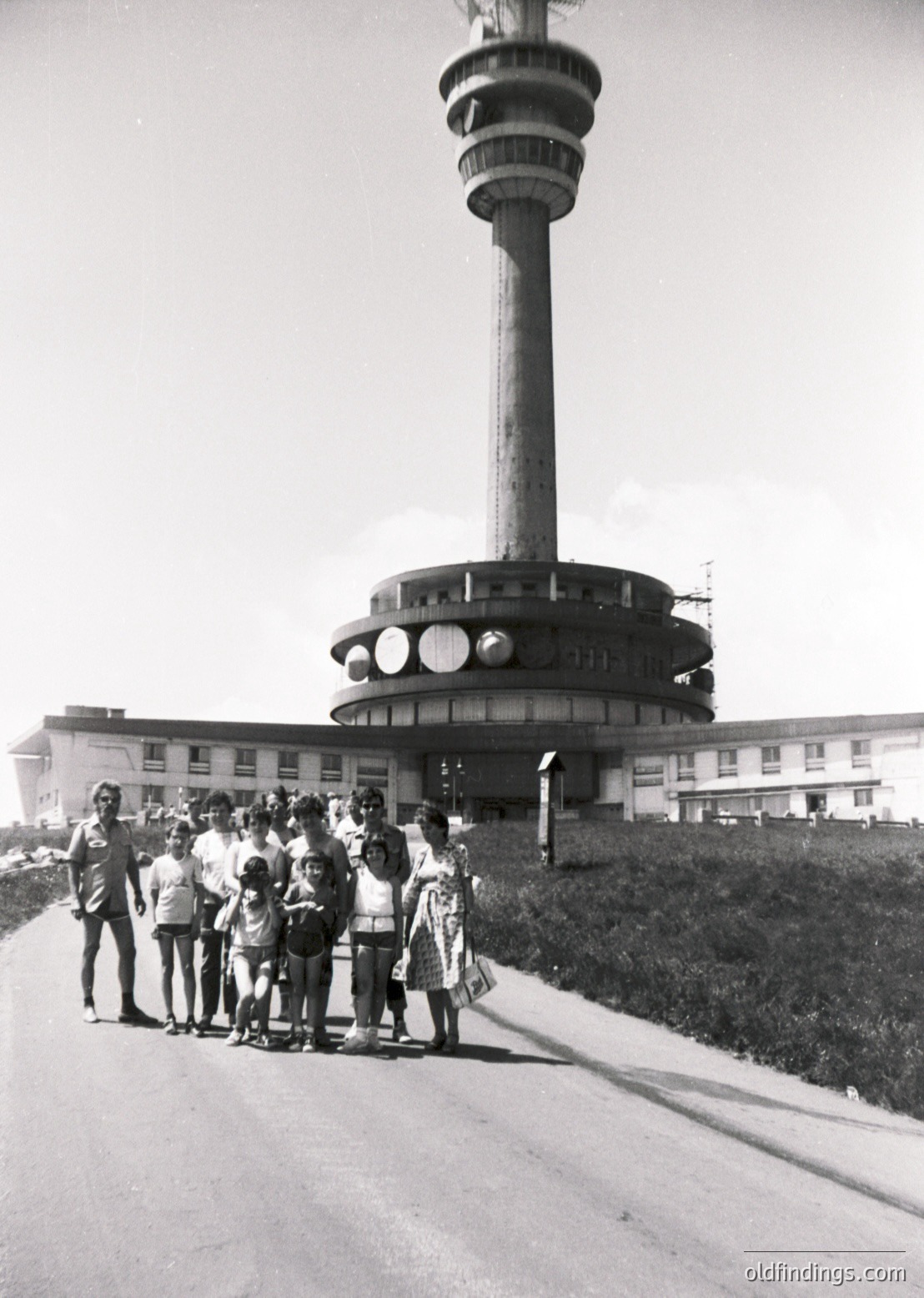 Iconic 1960s Soviet-era observation tower with Brutalist design, likely the *Teleferik Tower* in Varna, Bulgaria. Group of people—adults and children—pose in front of the tower’s circular base and elevated platform. Mid-century architecture with geometric, utilitarian styling.