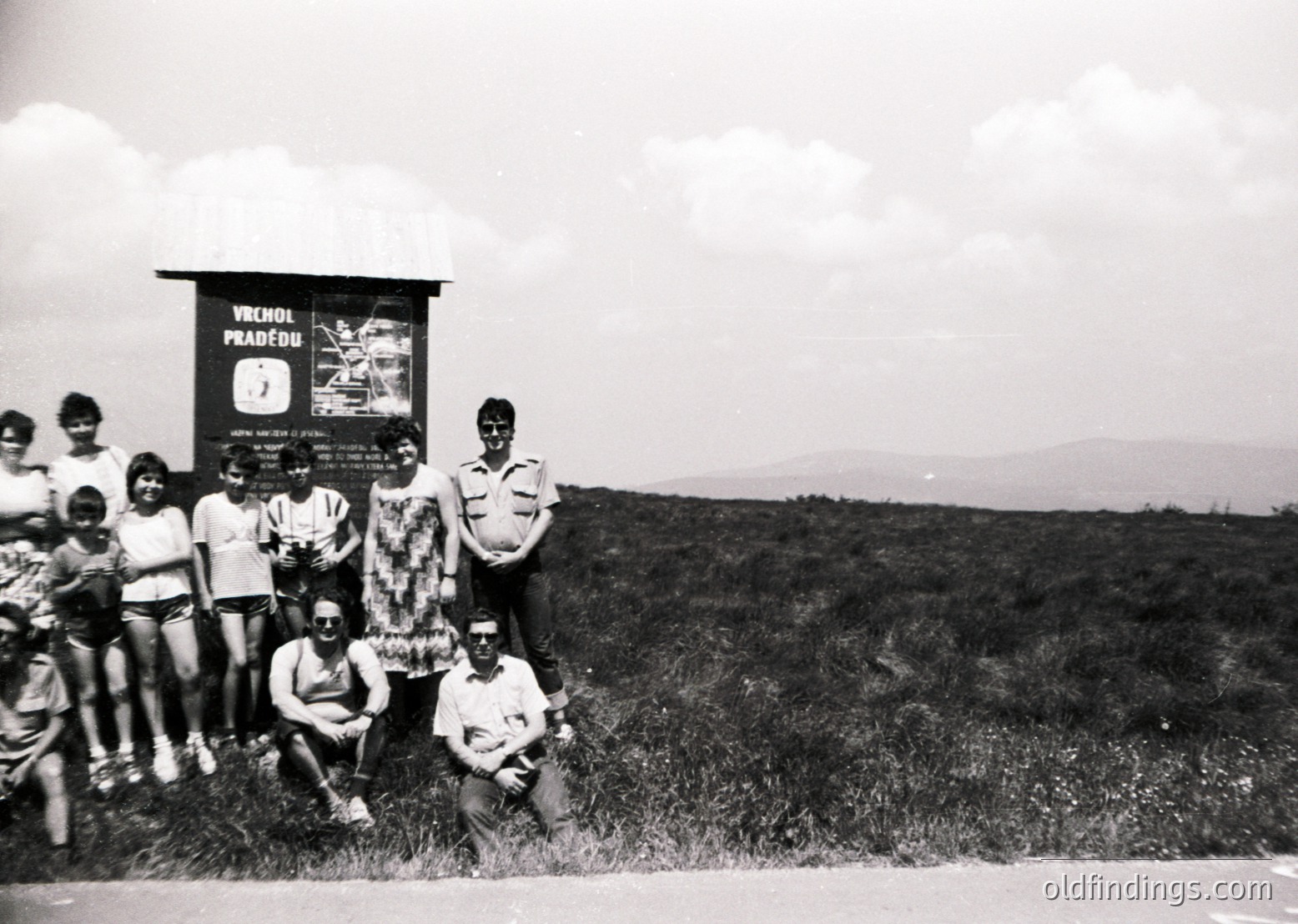 Black-and-white group photo at a mountain summit signpost reading "Vrchol Pradědu" (Praděd Peak, Czech Republic), 1960s–70s. Nine individuals pose with a scenic mountain backdrop. Sign includes hiking route details and a vintage TV poster. Casual summer attire (shorts, caps) suggests a recreational outing. ědu