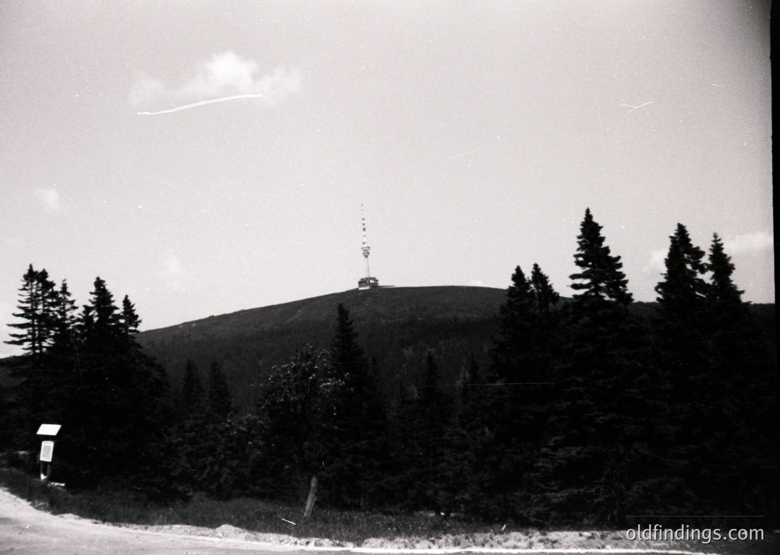 Black-and-white mountain landscape featuring a prominent radio tower atop a forested peak, surrounded by dense coniferous trees. Mid-20th century architectural structure with a triangular antenna. Clear skies with minimal cloud cover.