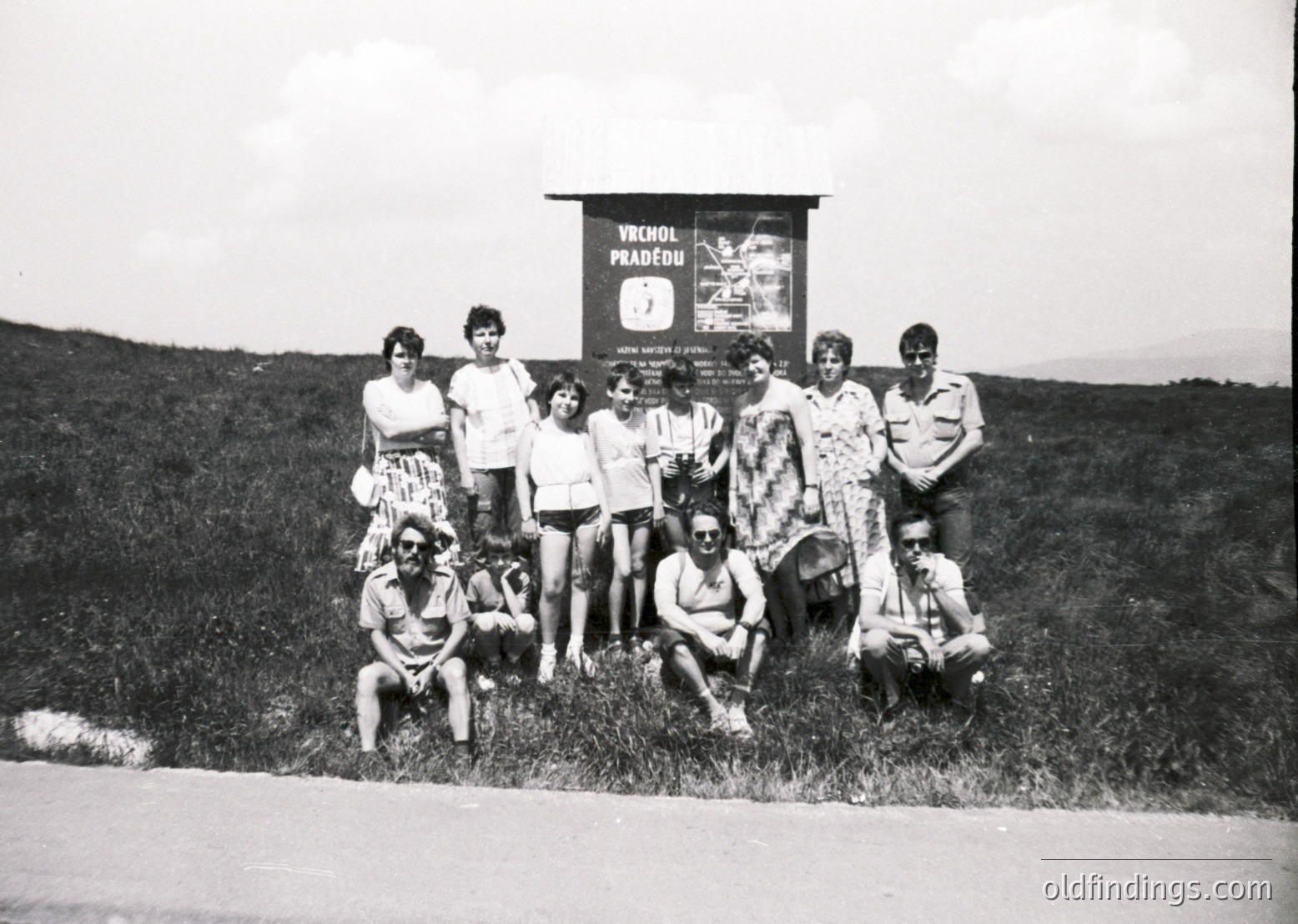 Group photo on grassy hillside beside road, 1960s Czechoslovakia. Sign reads "Vrchol Pradědu" (Praděd Peak). Nine standing, four kneeling, one child with dog. Casual summer attire: dresses, shorts, hats. ědu