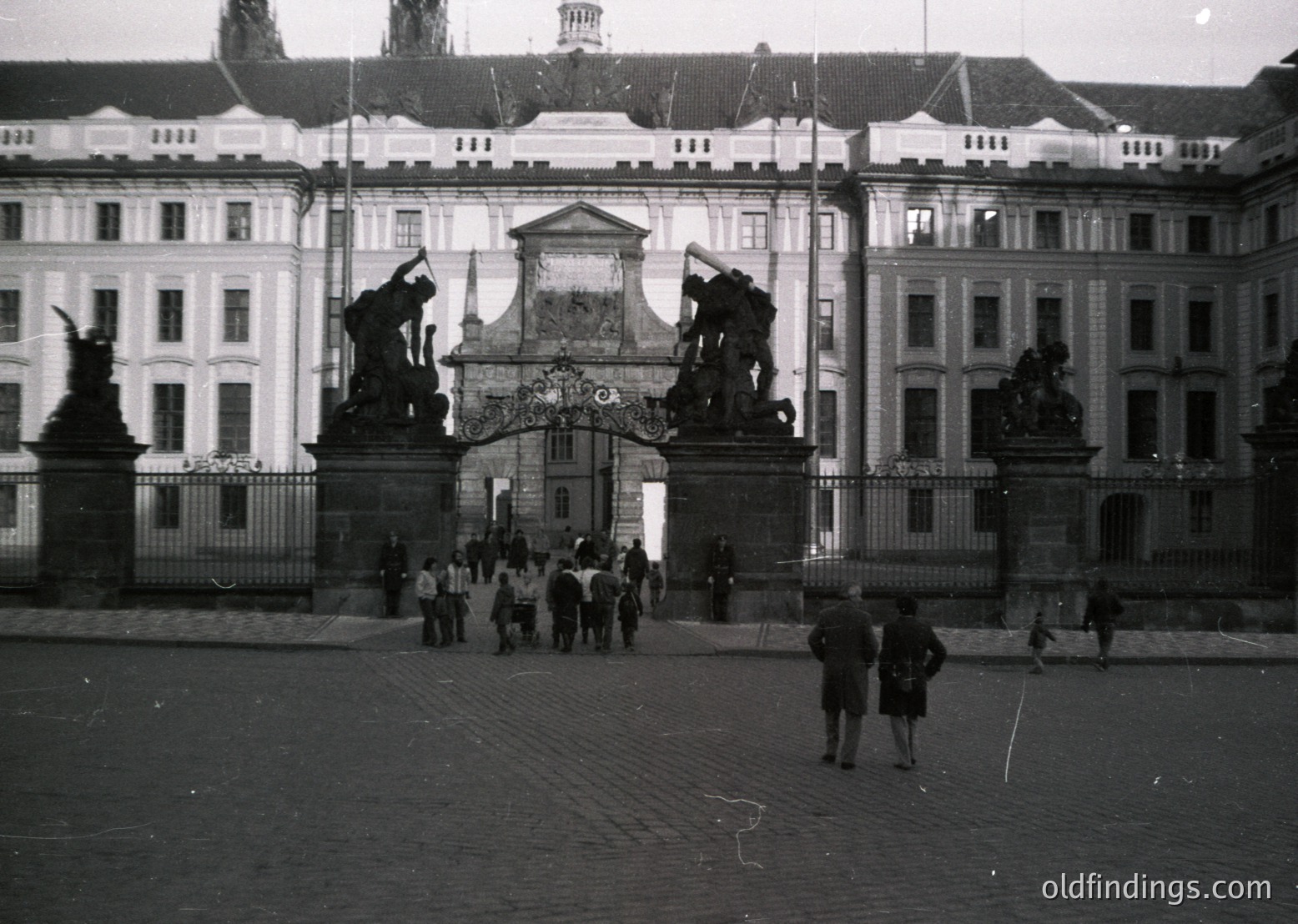 Neoclassical gatehouse with sculpted lions flanking an ornate archway, set against symmetrical palace wings. Mid-20th century black-and-white photo captures a courtyard with pedestrians. Architectural details include pilasters, balustrades, and a central pediment.