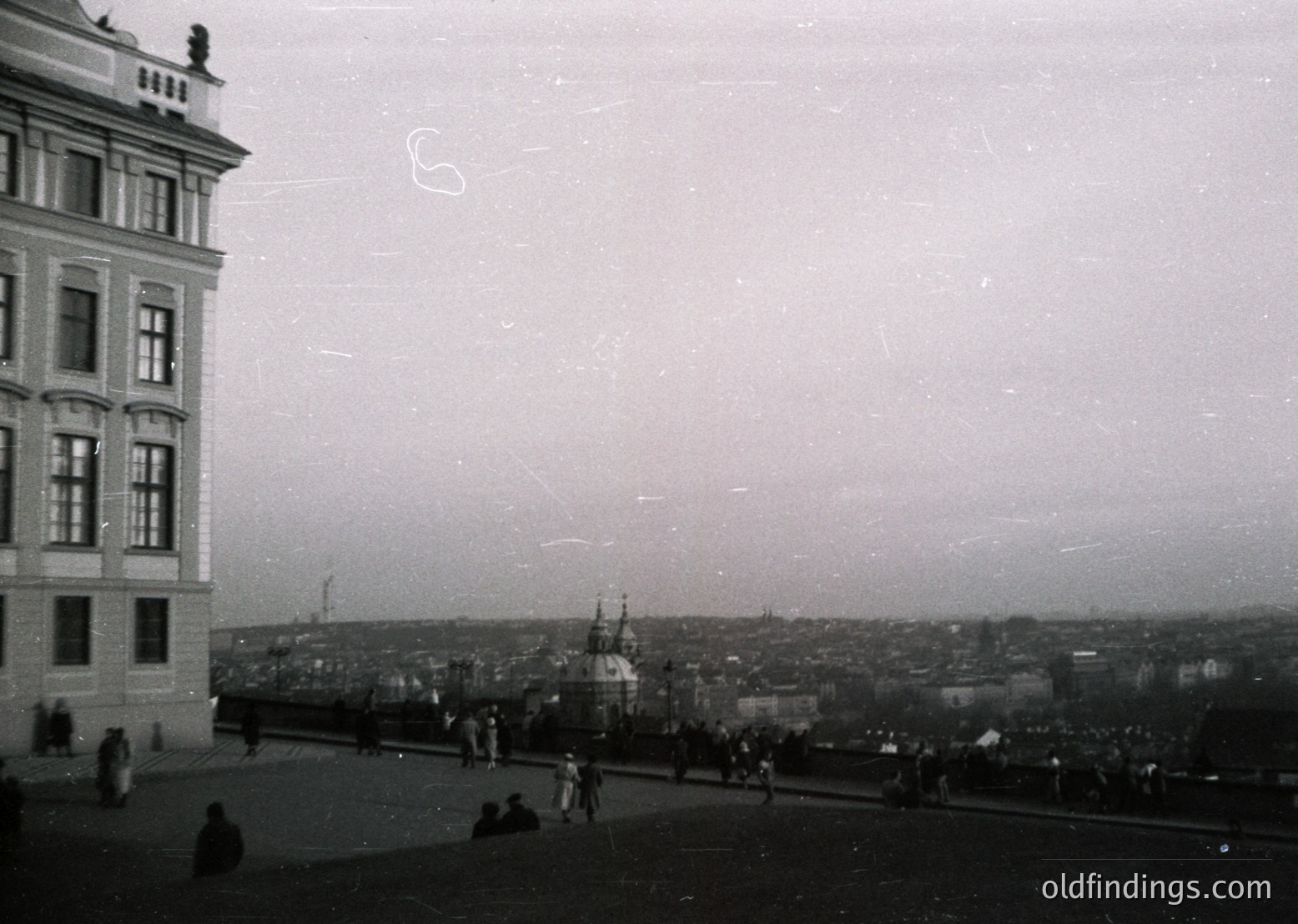Vintage black-and-white coastal cityscape featuring a grand, multi-story building with ornate architectural details. Prominent church spire and naval ships docked in the harbor below. Crowds of people strolling along a seaside promenade, suggesting a bustling seaside resort. Likely