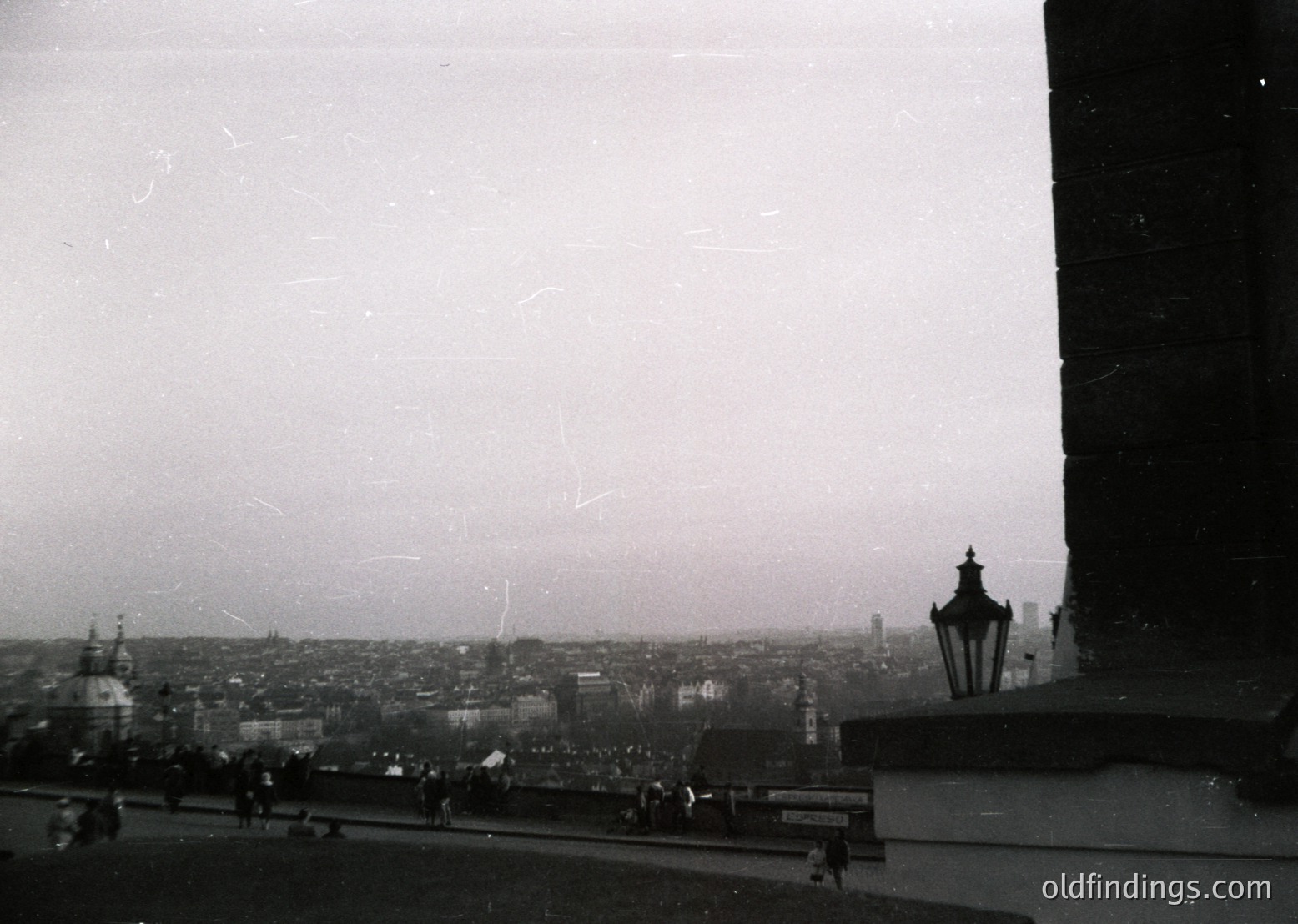Vintage black-and-white cityscape shot from an elevated viewpoint, likely a balcony or rooftop. Distinctive spire and ornate street lamp in foreground; hazy mid-20th century urban sprawl with low-rise buildings and visible tram tracks. Foggy atmosphere suggests early morning or industrial-era pollution.