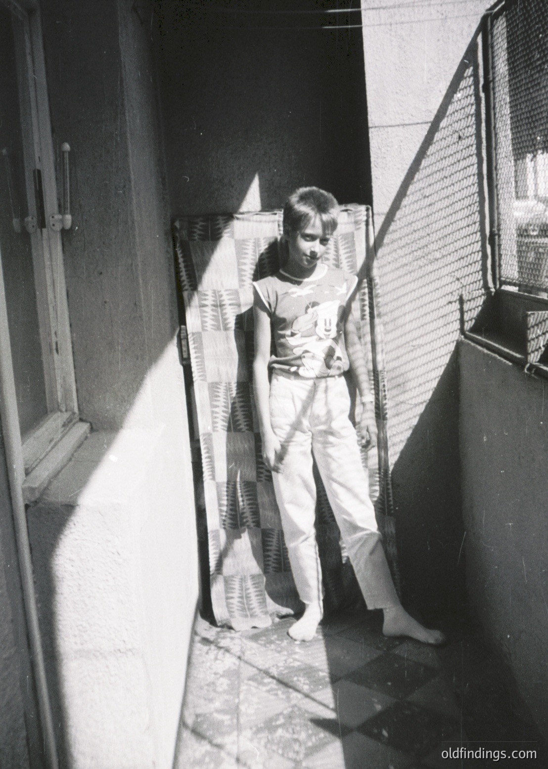 A young boy in 1960s-era prison uniform stands in a narrow, barred corridor, holding a wooden object. The institutional setting suggests a detention facility, likely Eastern Bloc (1960s–70s). Concrete walls, metal bars, and a single open door frame the scene.