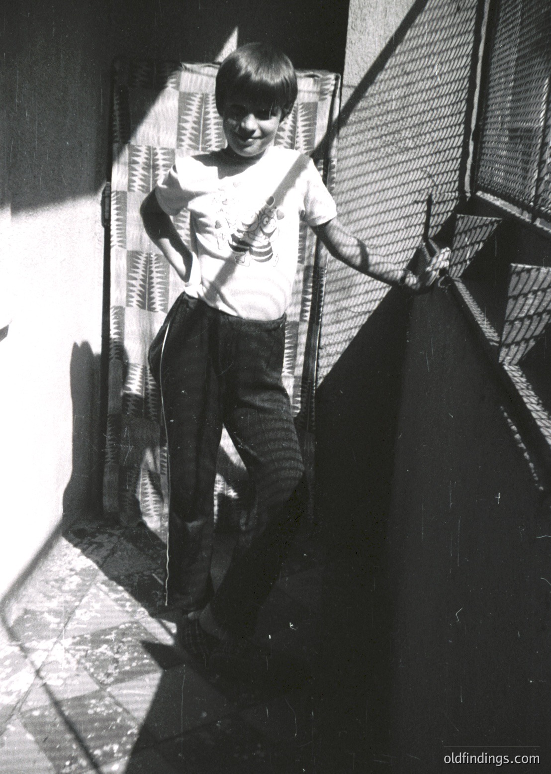 Young boy in 1960s-70s streetwear poses with acoustic guitar on urban staircase. Lighting suggests midday sun casting shadows.