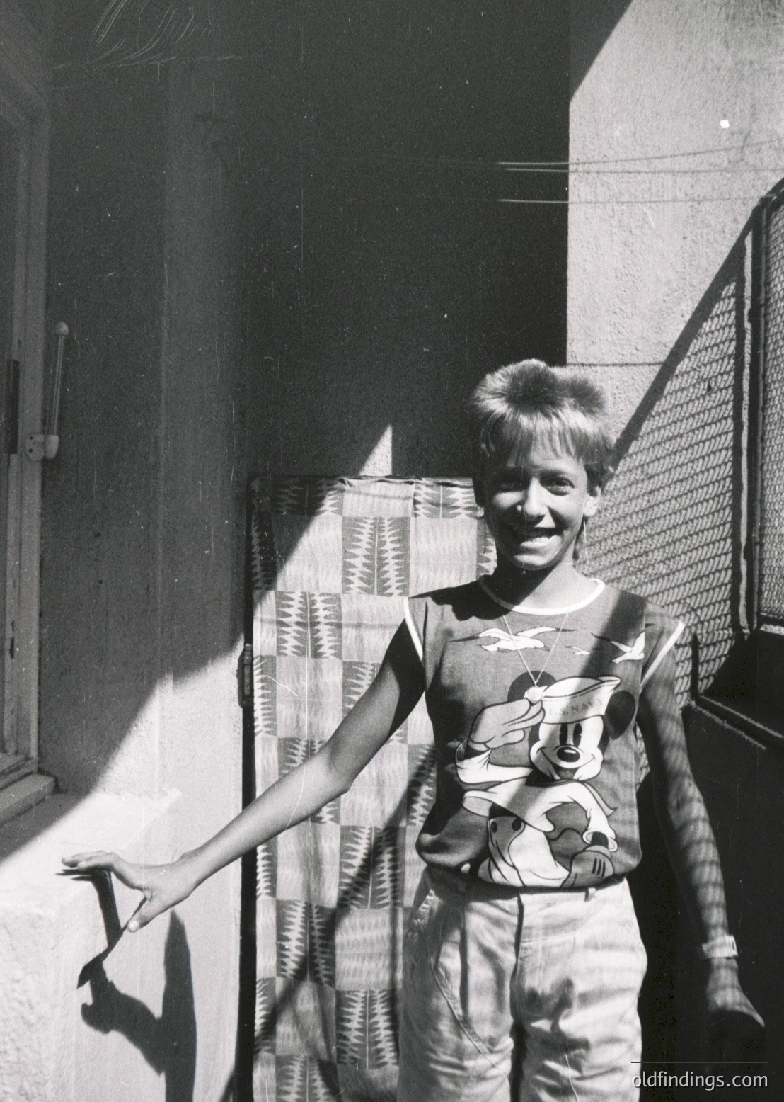 Mid-century boy in a graphic tank top and baggy pants poses outside a residential building, likely 1960s–1970s. Checkered curtain and metal fence detail suggest urban or suburban setting. Candid, sunlit portrait captures youthful energy.