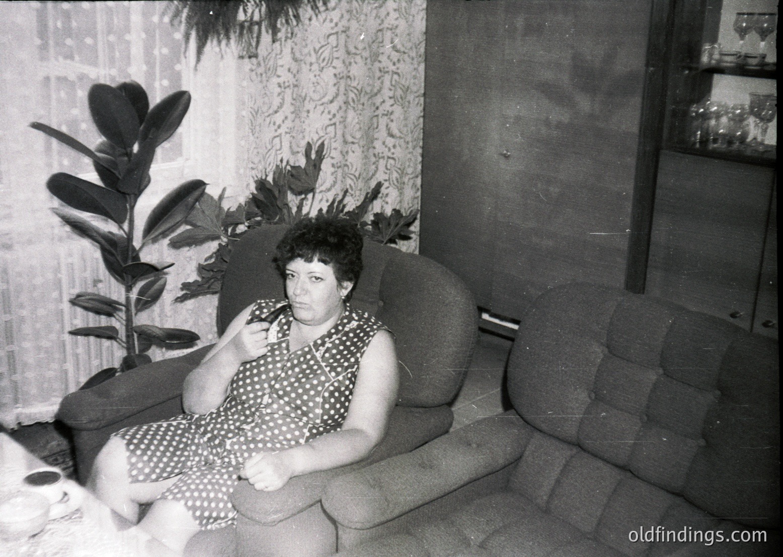 Vintage black-and-white portrait of a woman seated on a curved sofa in a mid-century interior, holding a cigarette. She wears a polka-dot dress and a headscarf. Surrounding greenery and patterned wallpaper suggest a 1960s–1970s European home.