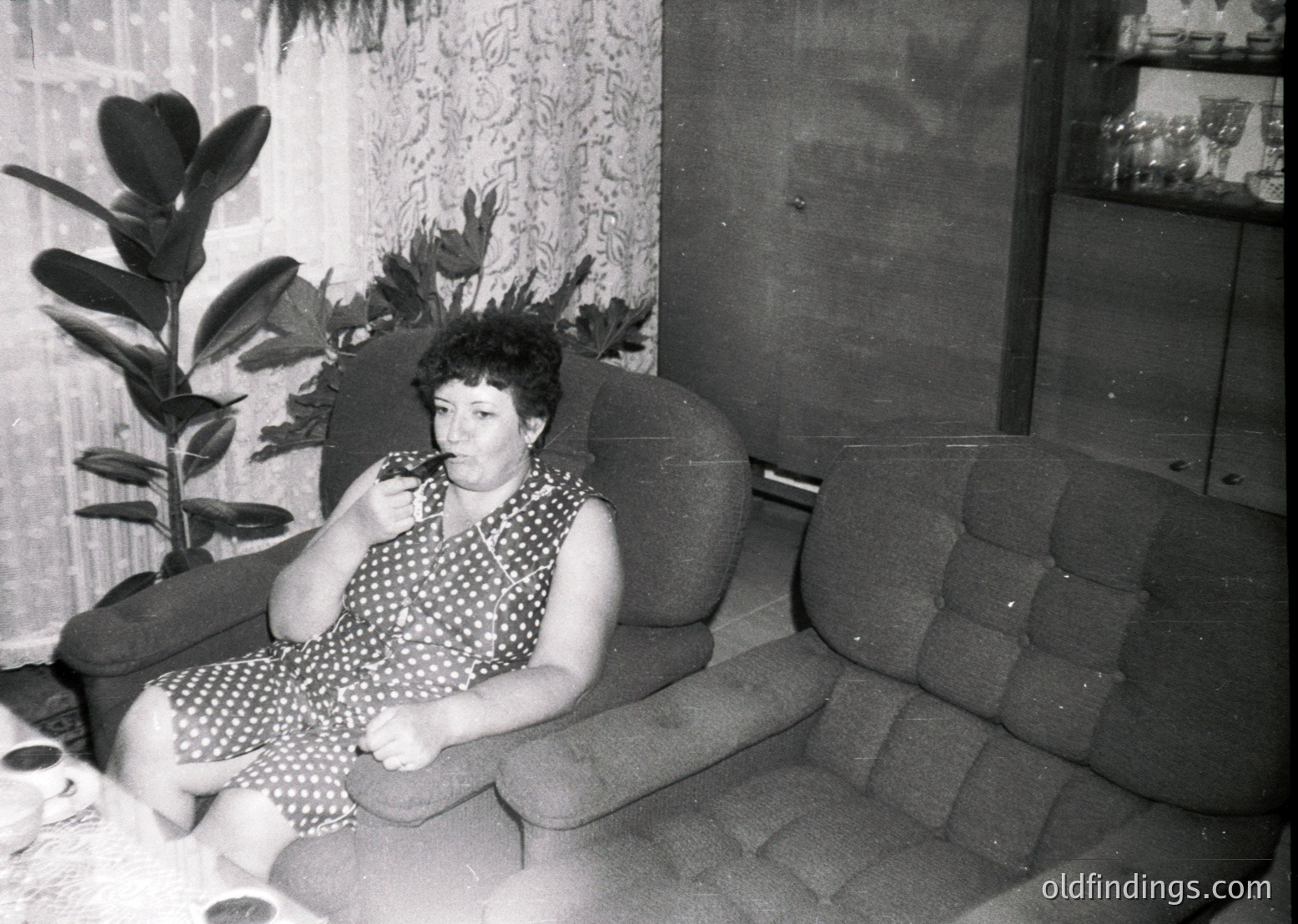 Mid-century indoor portrait of a woman in a polka-dot dress, seated on a tufted armchair beside a potted plant. Soft focus and grain suggest 1960s–1970s era. Reflective surfaces hint at a modestly furnished living space.