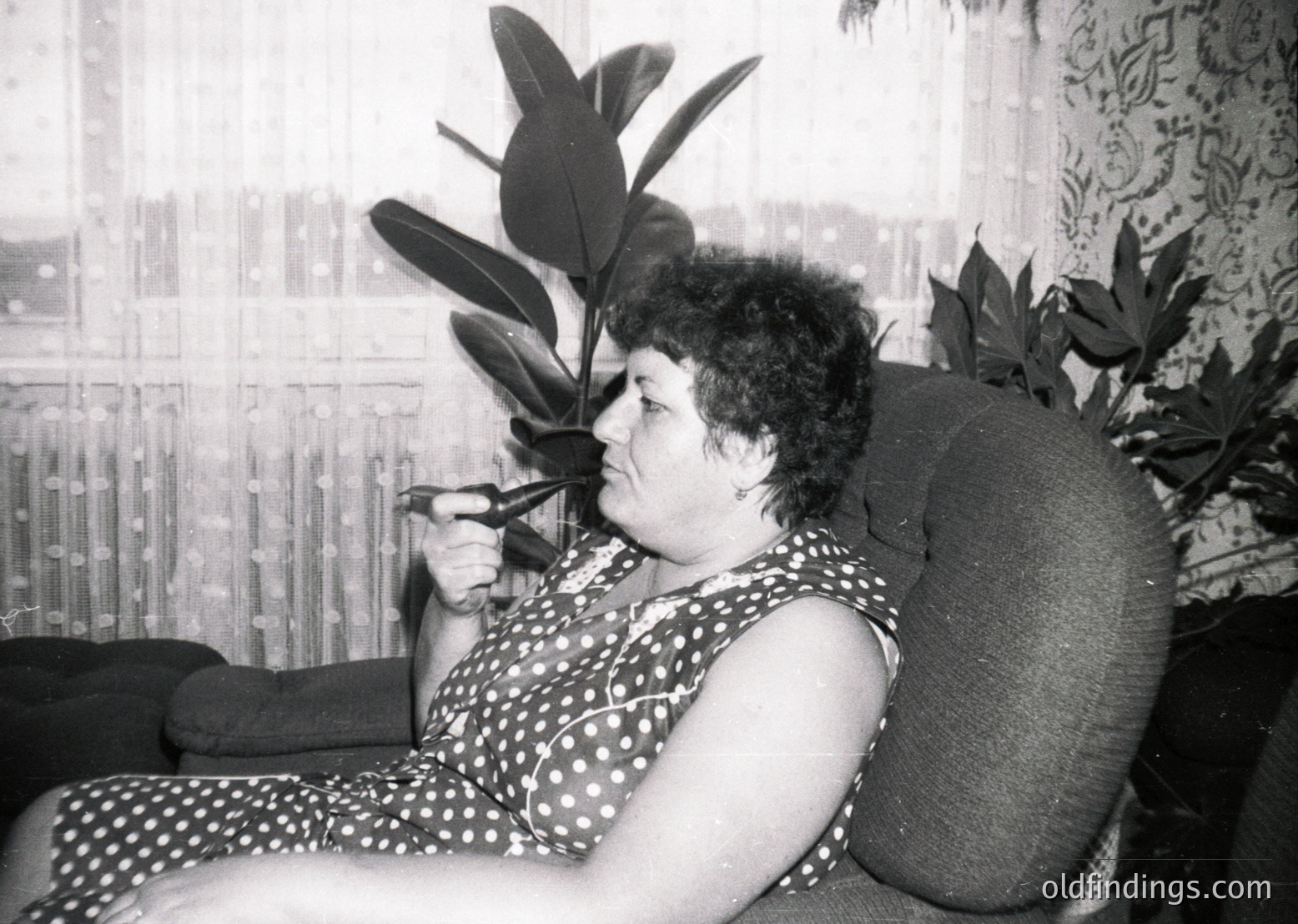 Mid-century indoor portrait of a woman in a polka-dot dress, holding a cigarette, seated on a tufted armchair beside a large leafy plant. Patterned curtains frame the window behind her. Likely 1960s–1970s domestic setting.