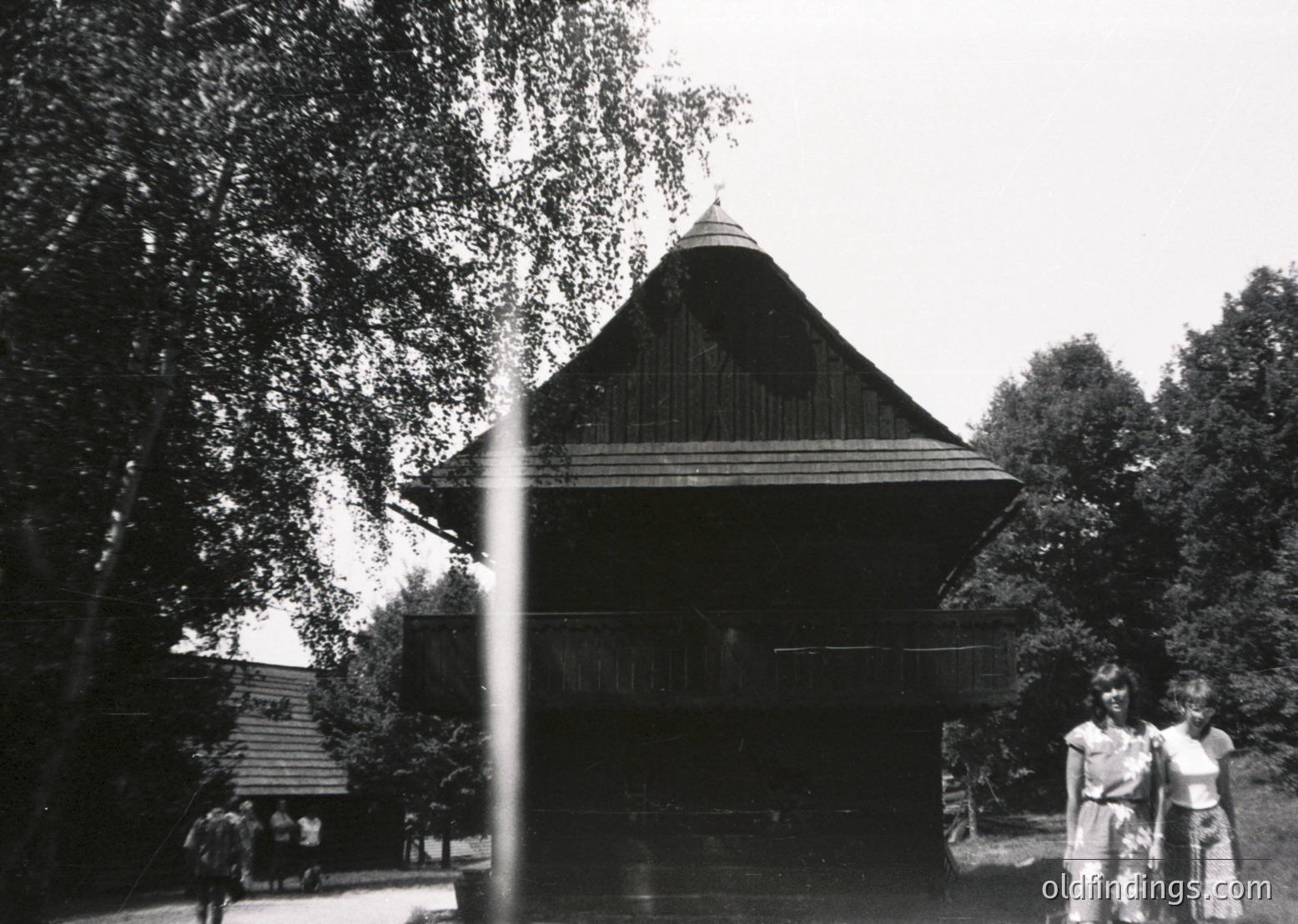 Traditional wooden pavilion with steep gabled roof, surrounded by lush greenery. Mid-20th century architectural style, likely Southeast Asian. Two figures in traditional attire pose near entrance, suggesting cultural heritage exhibit or festival setting.