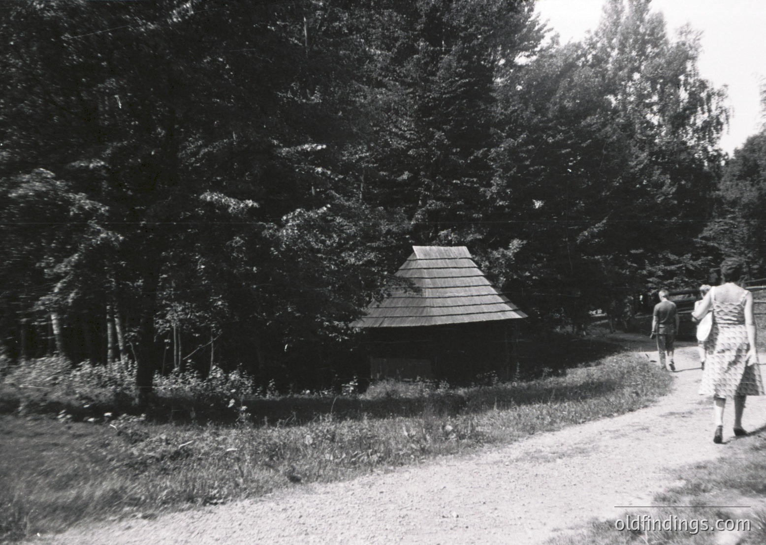 Mid-century wooden gazebo nestled in a forested area, surrounded by dense coniferous trees. Two figures in period attire (1950s-60s) walking along a gravel path. Rustic, serene landscape evokes mid-20th-century outdoor leisure.