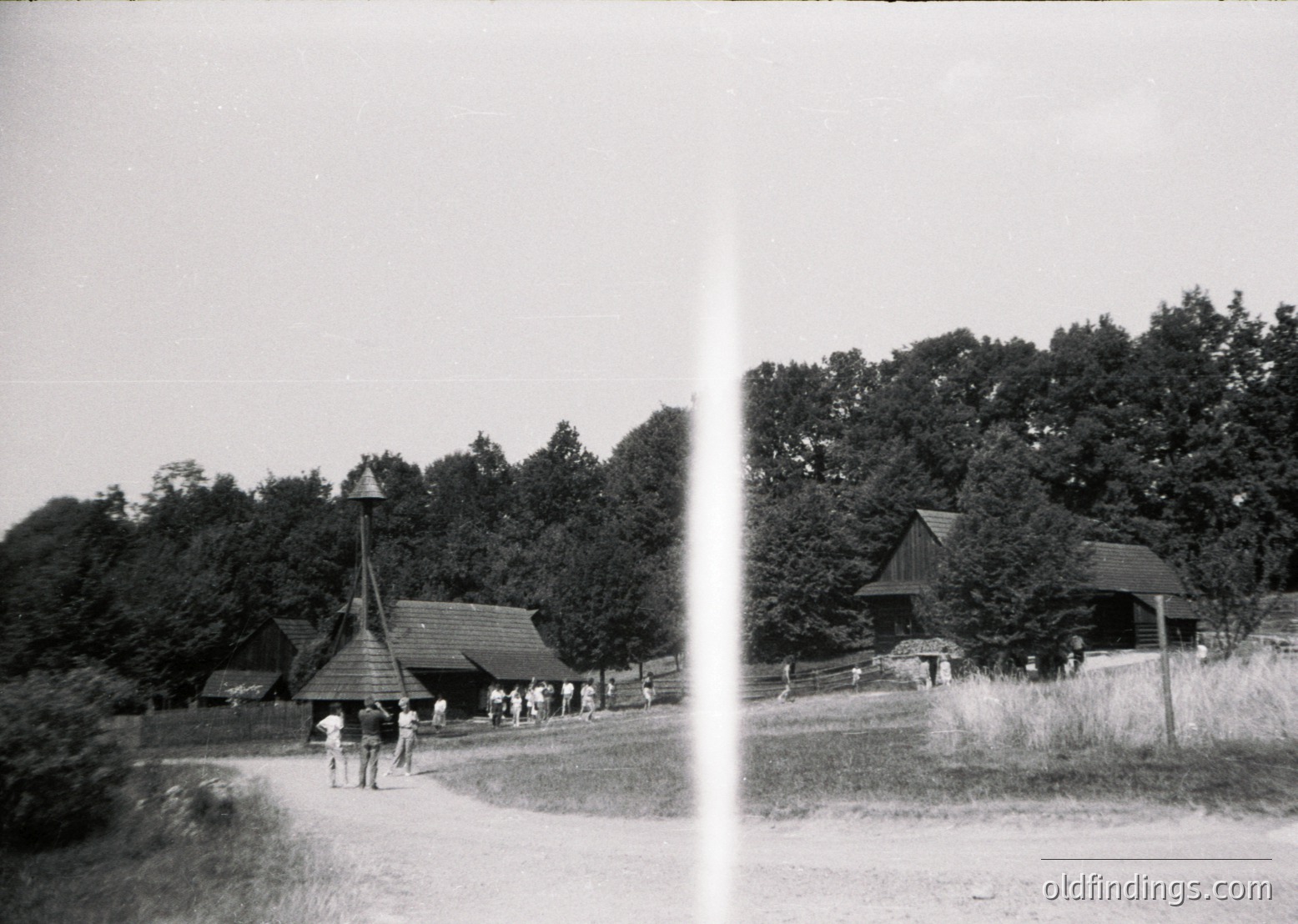 Black-and-white rural scene featuring a wooden pavilion with a conical roof, likely a folk architecture exhibit. Group of people in casual attire walking along a dirt path surrounded by dense forest. Open grassy area with a second wooden structure in background. Evokes mid-20th century Eastern European village life.