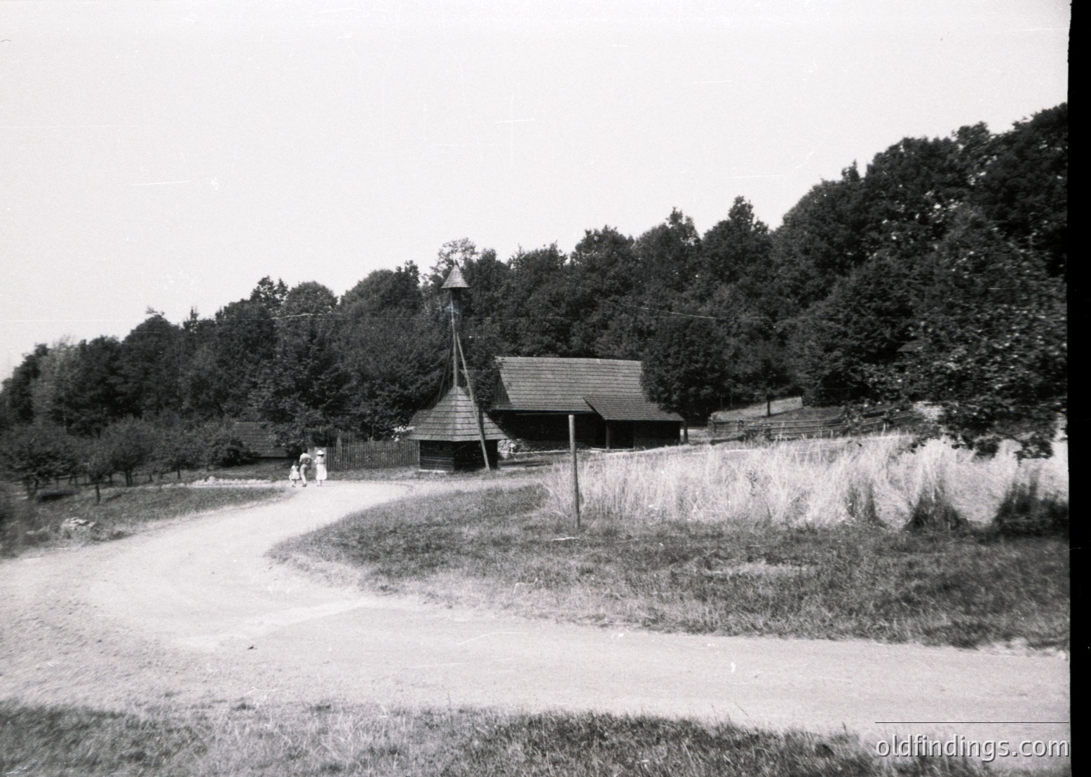 Rural countryside scene featuring a wooden barn with a steep gable roof and a small steeple, surrounded by dense forest. Two figures walk along a winding dirt road bordered by hay bales. Mid-20th century farmstead, likely Eastern Europe.