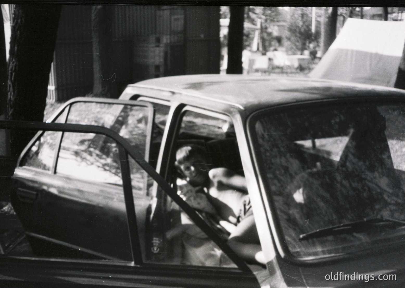 Classic mid-century sedan parked beside a tree-lined sidewalk, likely Eastern European . Driver’s side door open, revealing a man in dark clothing, hand near steering wheel. Urban street scene with blurred background indicating motion. Potential vintage automotive or urban history reference.
