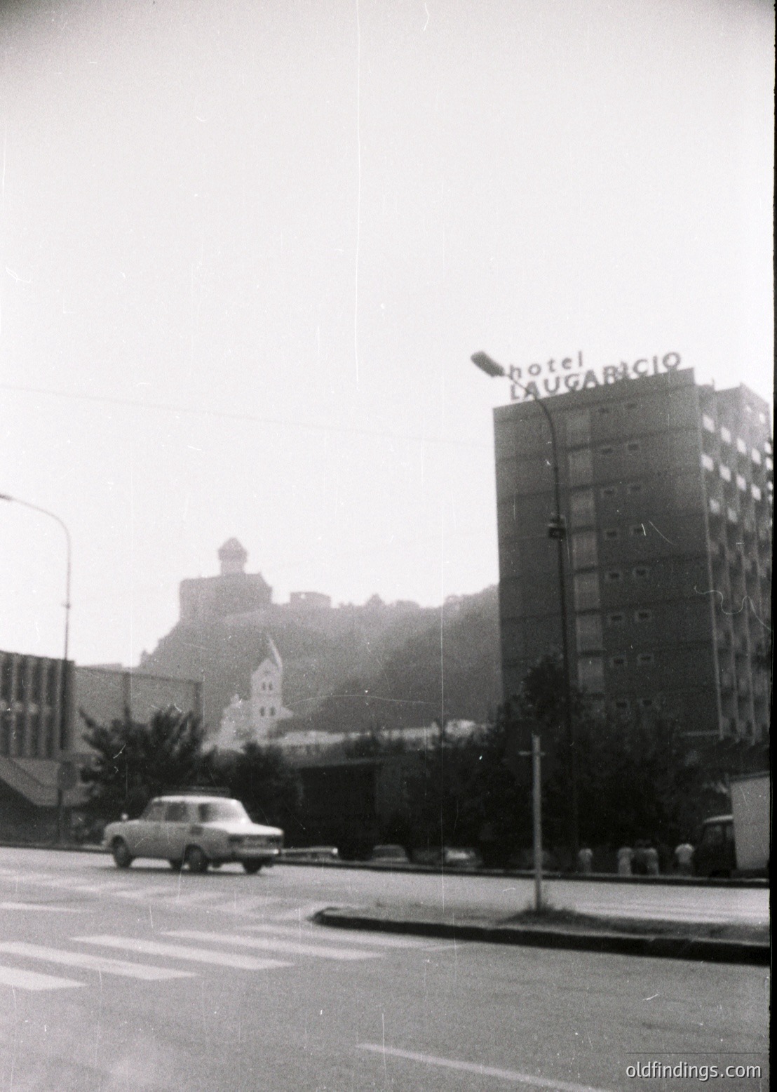 Black-and-white urban street scene featuring **Hotel Dugaricio** (1960s-70s style signage) in foreground, likely **Sofia, Bulgaria**. Mid-century concrete high-rise with vertical window bands dominates. Classic **Volga car** (1960s-70s) driving past, with **pedestrian crosswalk** and sparse greenery. Historic **church tower** visible in background.