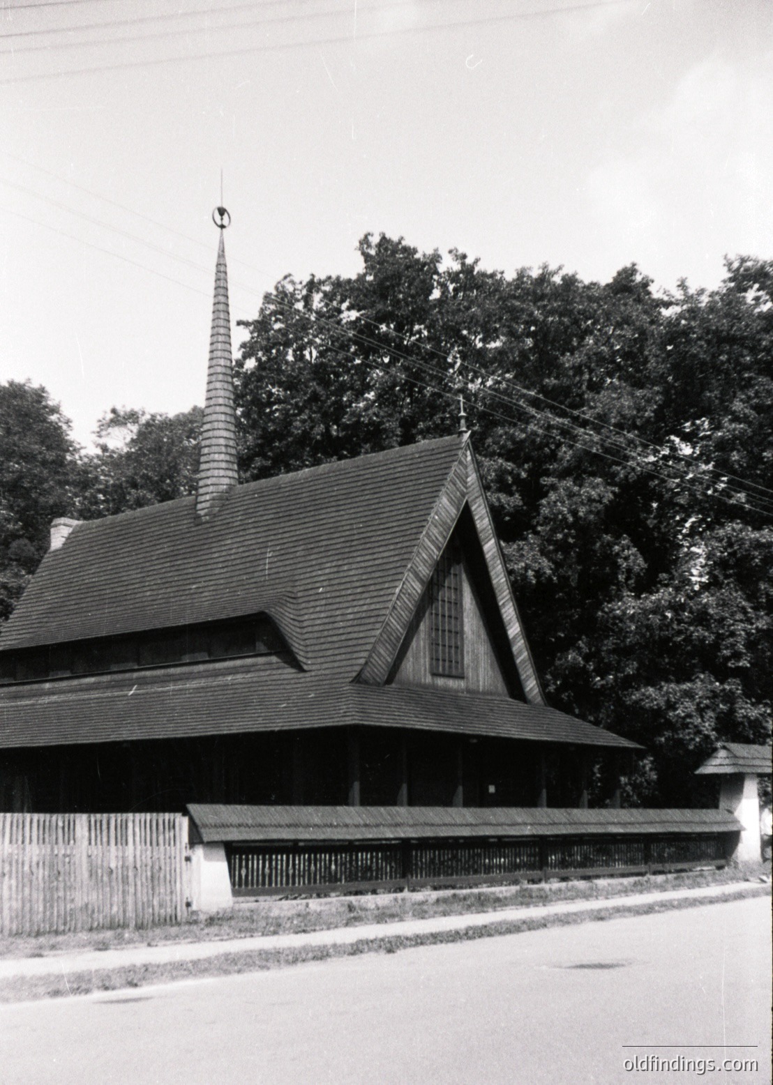 Wooden church with steep gable roof and tall, slender spire, likely Eastern European architecture. Rustic fence and mature trees frame the structure, suggesting rural or suburban setting.
