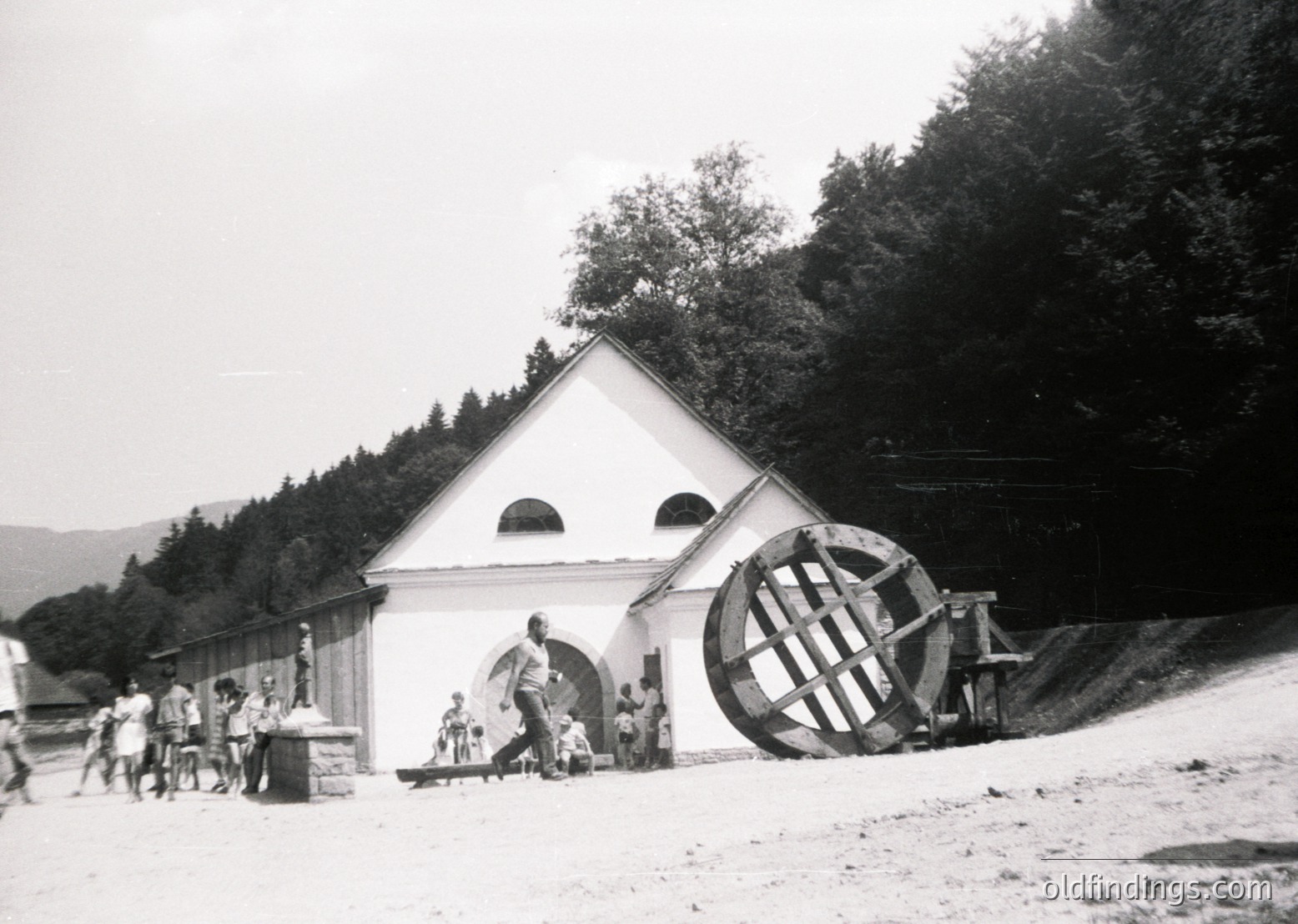 Historic waterwheel-powered mill in a rural alpine setting, likely 1940s-1950s. White triangular-roofed structure with arched windows and a large wooden wheel beside a stone pathway. Group of people, including adults and children, gathered around. Surrounded by dense forest and rolling hills.