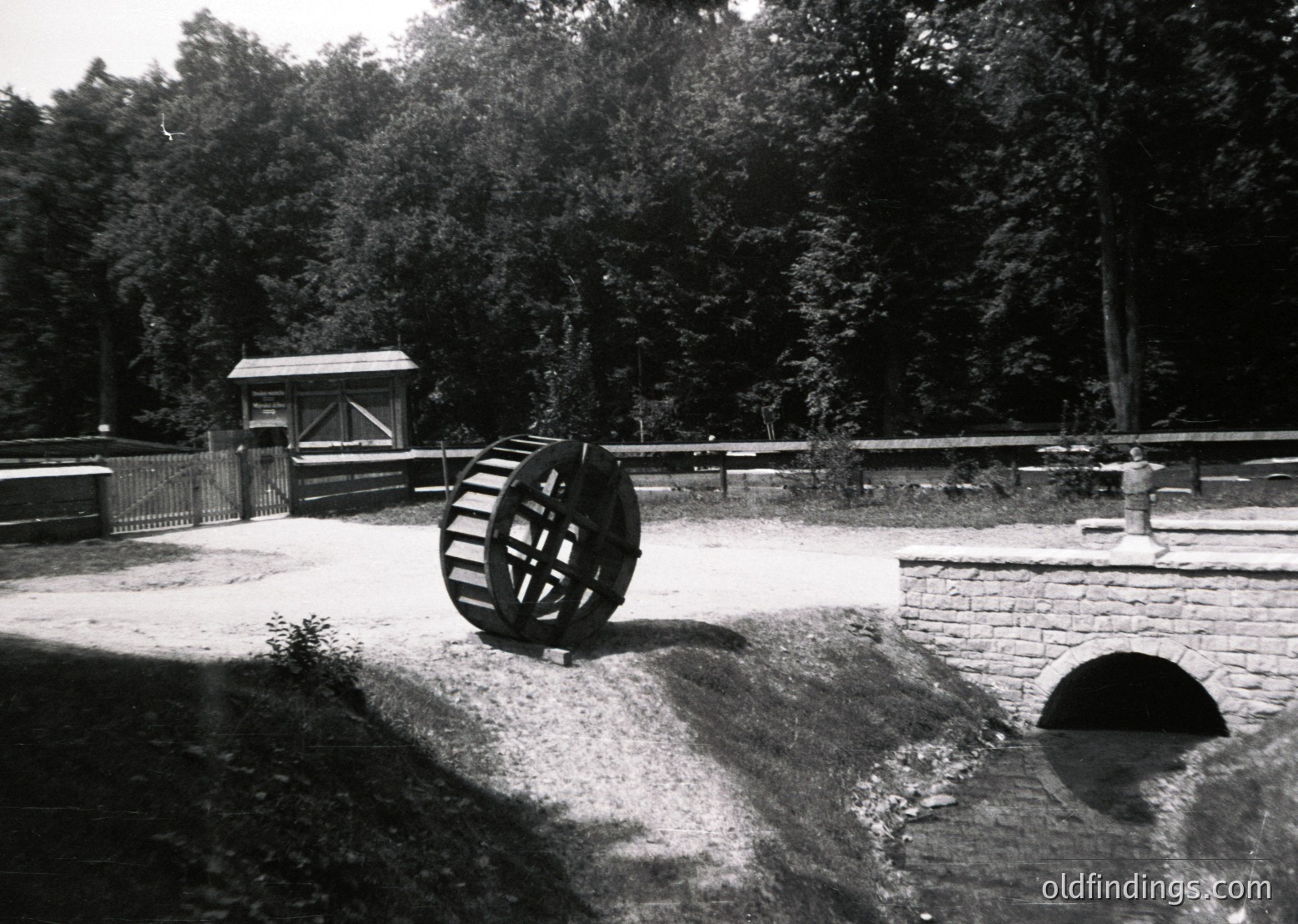 Historic waterwheel in a rural setting, likely part of an old mill or irrigation system. The brick-lined channel and wooden guardrail suggest 19th–early 20th century engineering. Dense forest backdrop indicates a temperate climate. Ideal for industrial heritage or vintage design references.