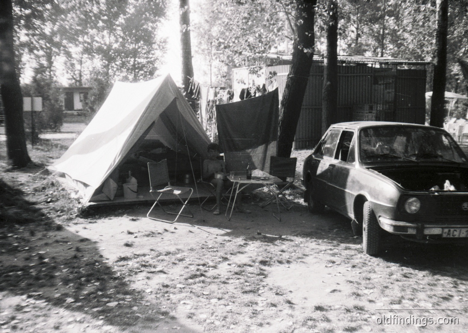 Vintage black-and-white camping scene featuring a canvas tent, folding chairs, and a mid-century sedan parked on a dirt path. Surrounding trees and a gated building suggest a park or recreational area. Likely mid-20th century (1950s–1970s).