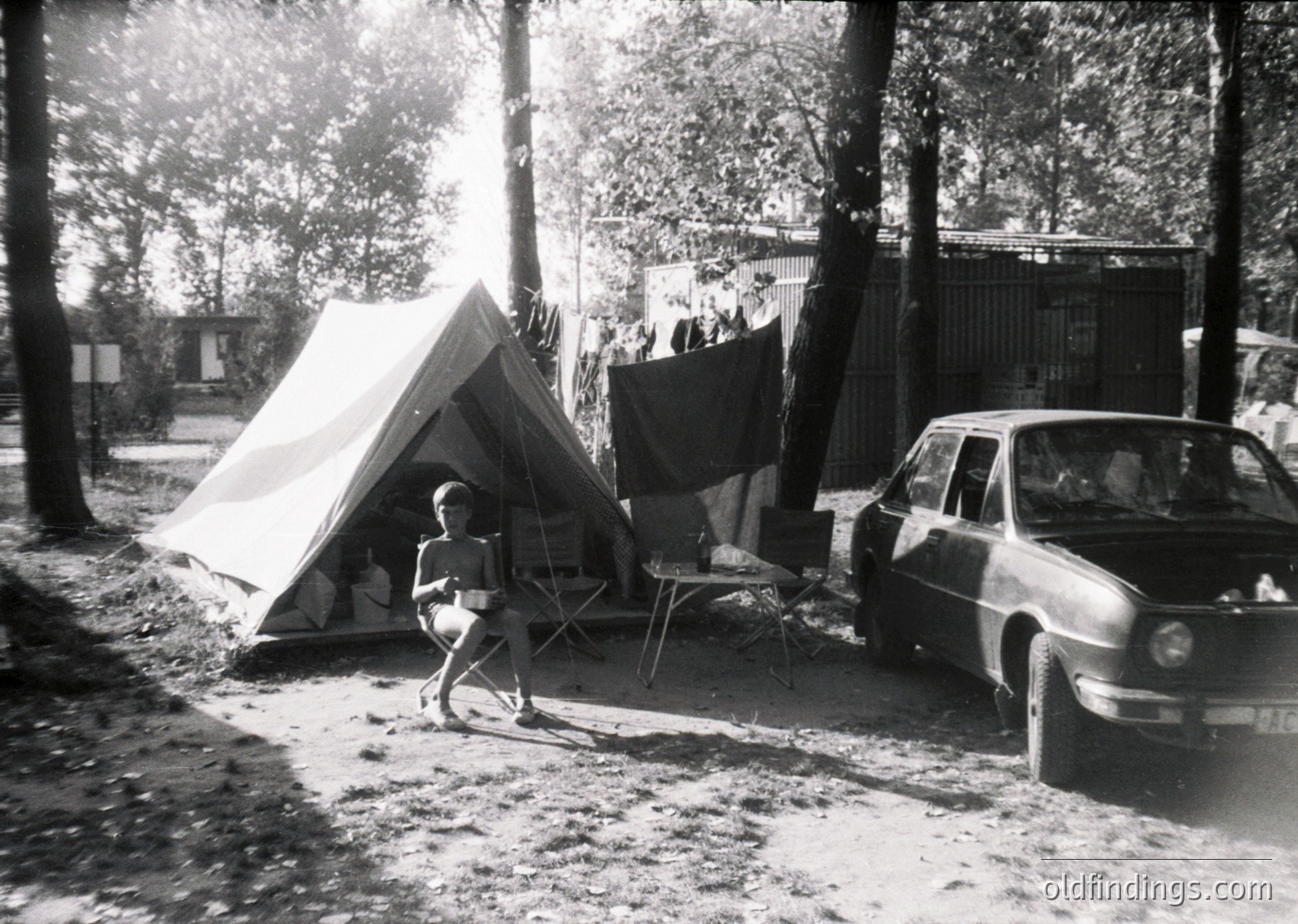 Mid-century camping scene with vintage tent and classic car. Child in shorts and shirt stands near a canvas tent pitched on a dirt path, surrounded by trees. A 1960s-era station wagon parked nearby. Laundry hung on a fence in background suggests a temporary residential setup. Evokes post-war outdoor living and family recreation.