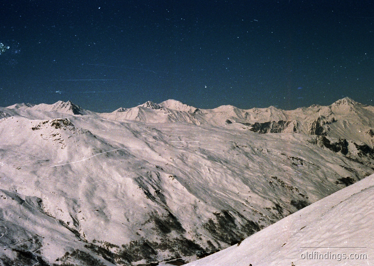 Stark alpine peaks under a star-studded night sky, showcasing jagged ridges and snow-covered slopes. The contrast of light and shadow highlights rugged terrain. Likely captured with vintage film, evoking mid-20th century exploration aesthetics.