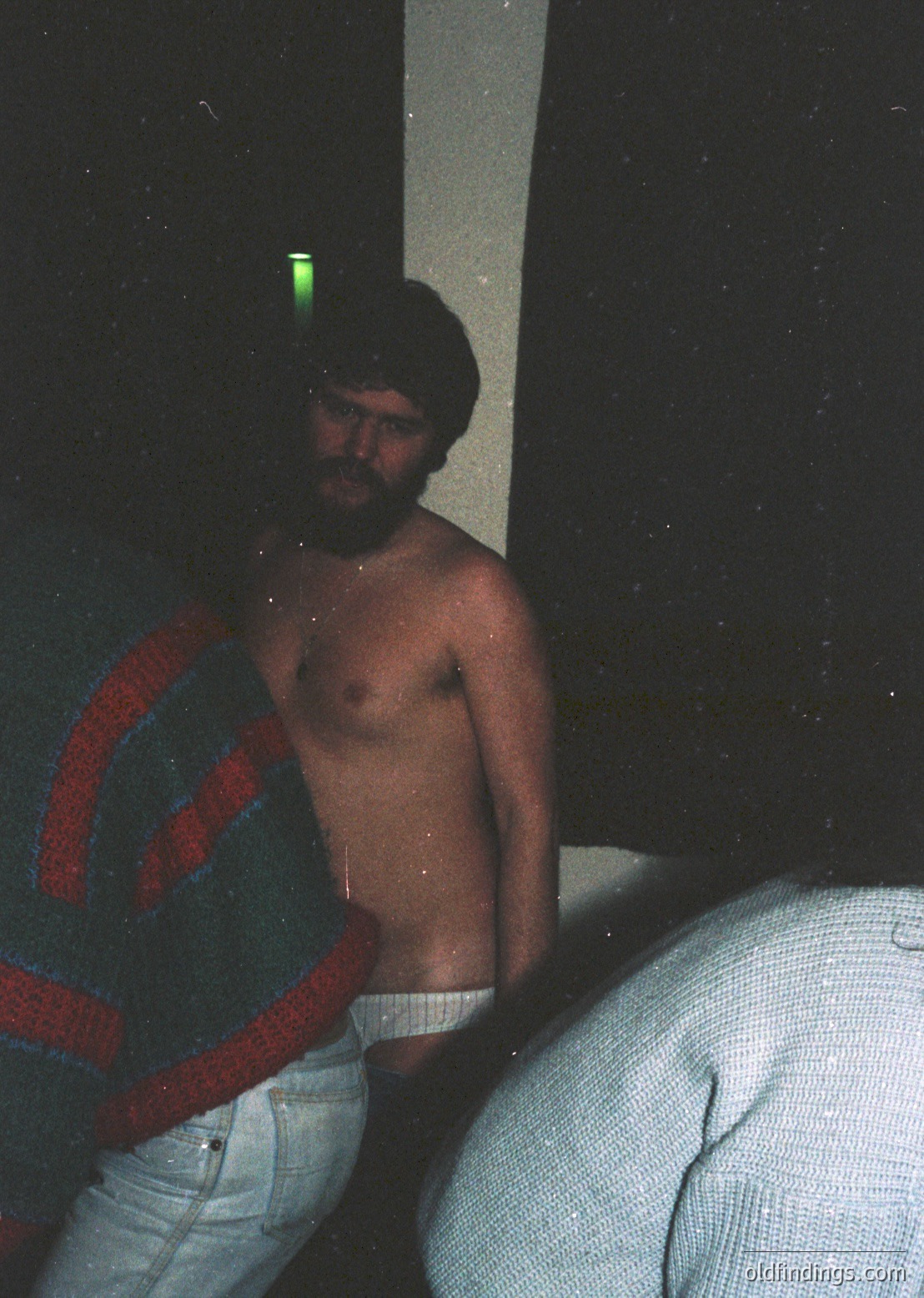 Vintage Polaroid-style photo of a shirtless man in low-light, holding a green fluorescent object. He wears light-colored pants with a red-and-blue striped sweater draped over his shoulder. Blurred background suggests motion or shallow depth of field. Likely late 20th century, possibly 1970s–1980s.