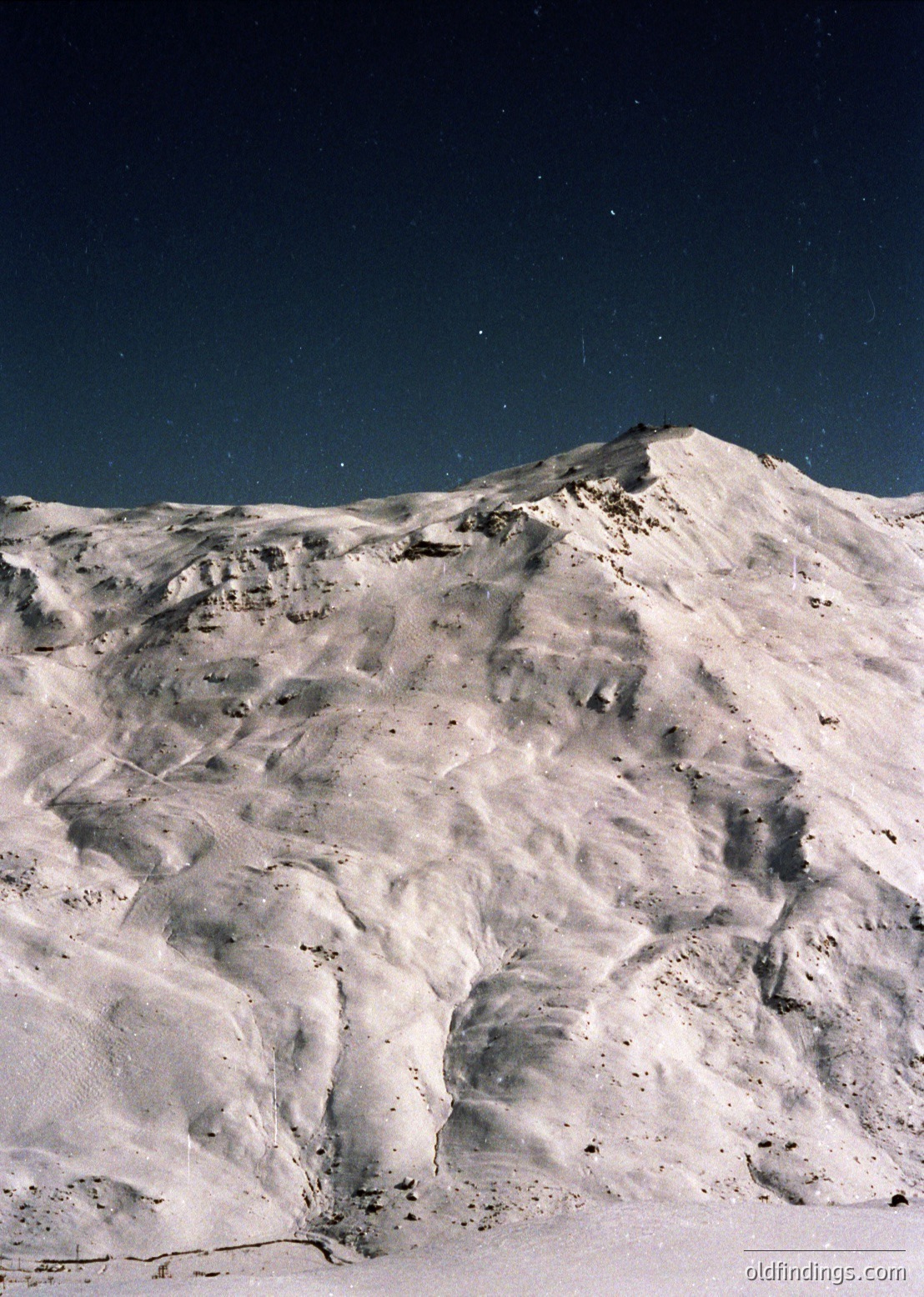 Snow-covered alpine peak under starry night sky, showcasing rugged terrain and dramatic lighting.