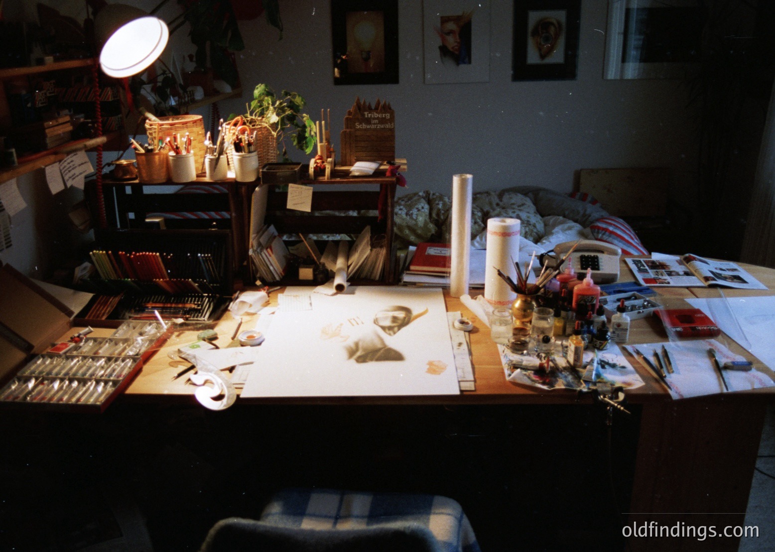 Cluttered artist’s workspace with vintage tools and supplies. Wooden desk bathed in natural light, featuring paintbrushes, sculpting tools, and a plaster cast. Shelves hold books, framed art, and a potted plant. Wall-mounted lamp illuminates the scene. Likely mid-20th century studio setting.