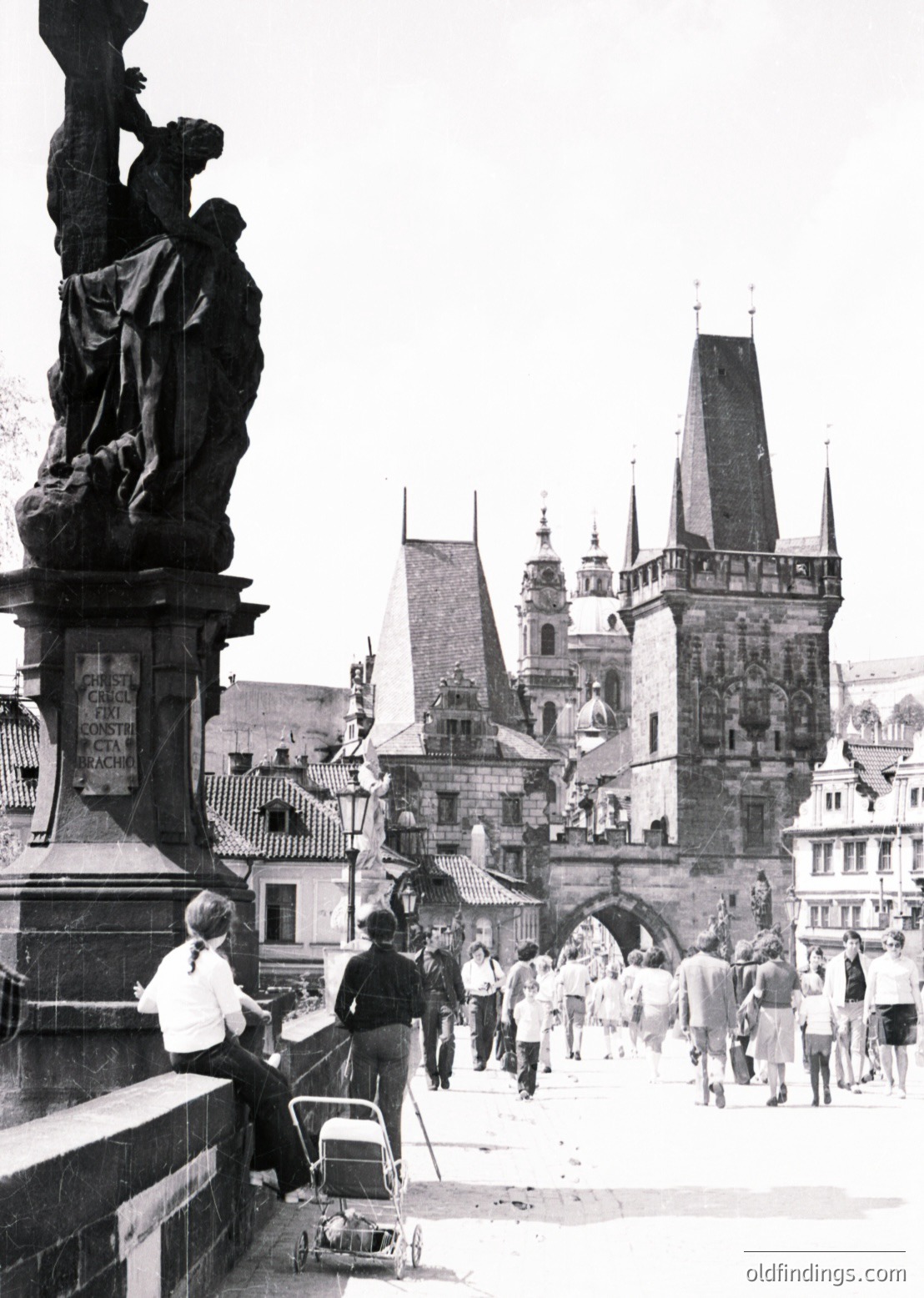 Black-and-white shot of Prague’s Charles Bridge (14th century) featuring Gothic-style statues and medieval architecture. Crowds in 1960s-era clothing walk along the bridge, while a musician plays near a statue. The Old Town Tower and spires of Prague Castle loom in the background.