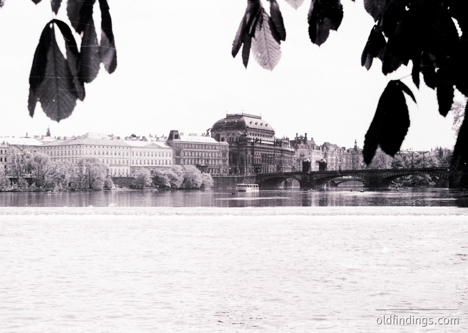 Mid-20th century urban riverscape: grand neoclassical buildings flank a bridge over calm water, framed by autumn leaves. Likely European cityscape,