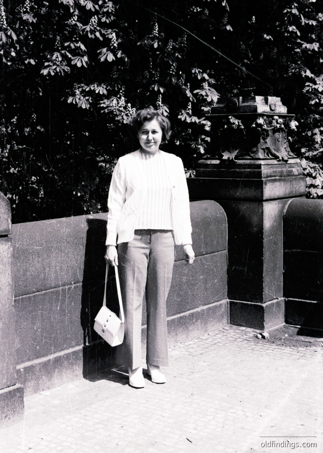 Mid-century woman in a light sweater and wide-leg trousers poses outdoors beside a stone wall and ornate lamp post. Classic 1960s–1970s fashion with structured handbag. Urban park or garden setting.