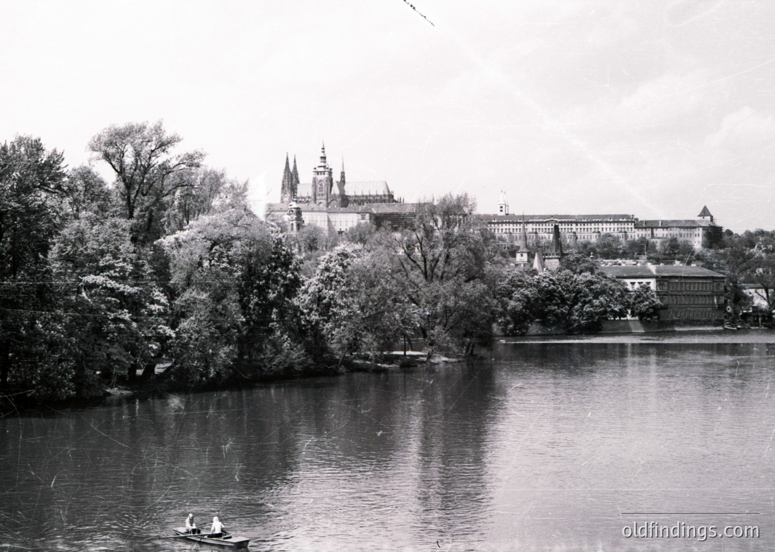 Historic black-and-white view of Prague Castle and the Vltava River, framed by autumn foliage. Gothic spires dominate the skyline, with classical palace wings extending along the hillside. Mid-20th century urban landscape with minimal modern interference.