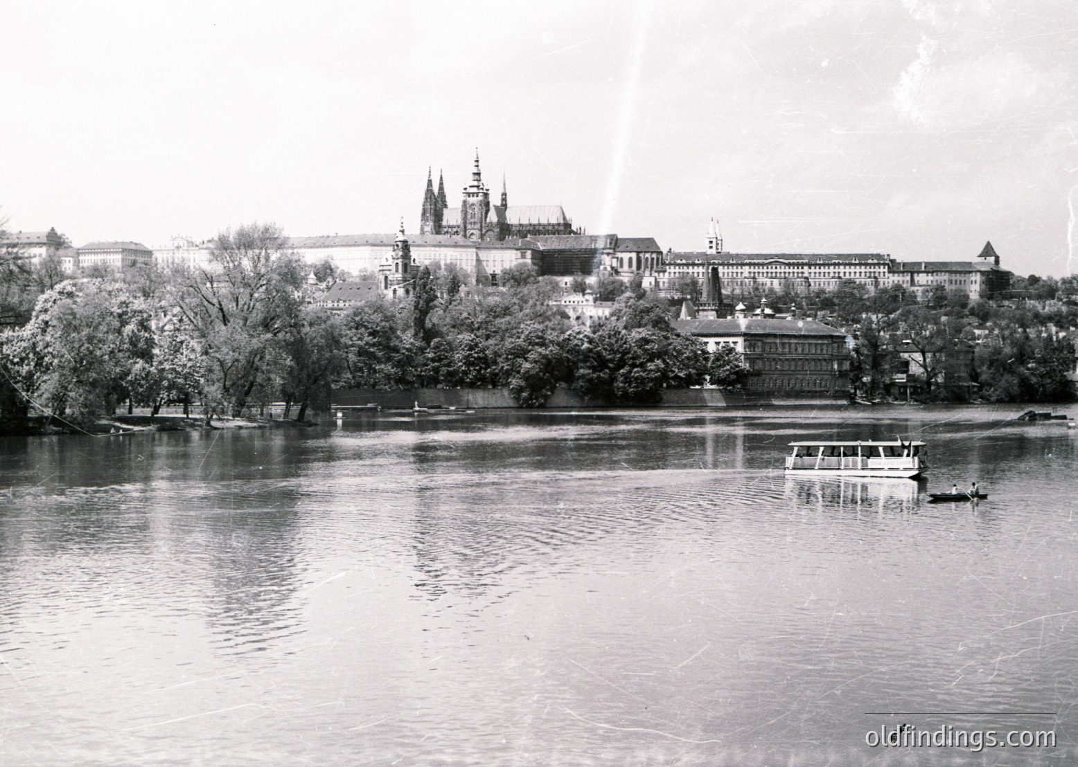 Vistula River panorama of the Wawel Royal Castle and Barbican in Kraków, Poland, mid-20th century. Gothic spires dominate the skyline, with a small boat navigating the river below. Lush greenery frames the historic fortress, reflecting its cultural significance.