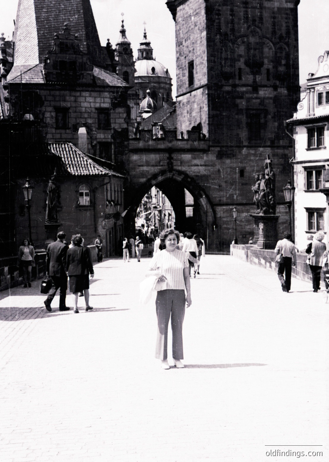 Mid-20th century black-and-white shot of Prague’s Charles Bridge, featuring Gothic arches, domed towers, and historic lanterns. Woman in striped blouse and skirt poses near the bridge’s central arch, surrounded by pedestrians in 1950s-60s attire. Iconic meets vibes.