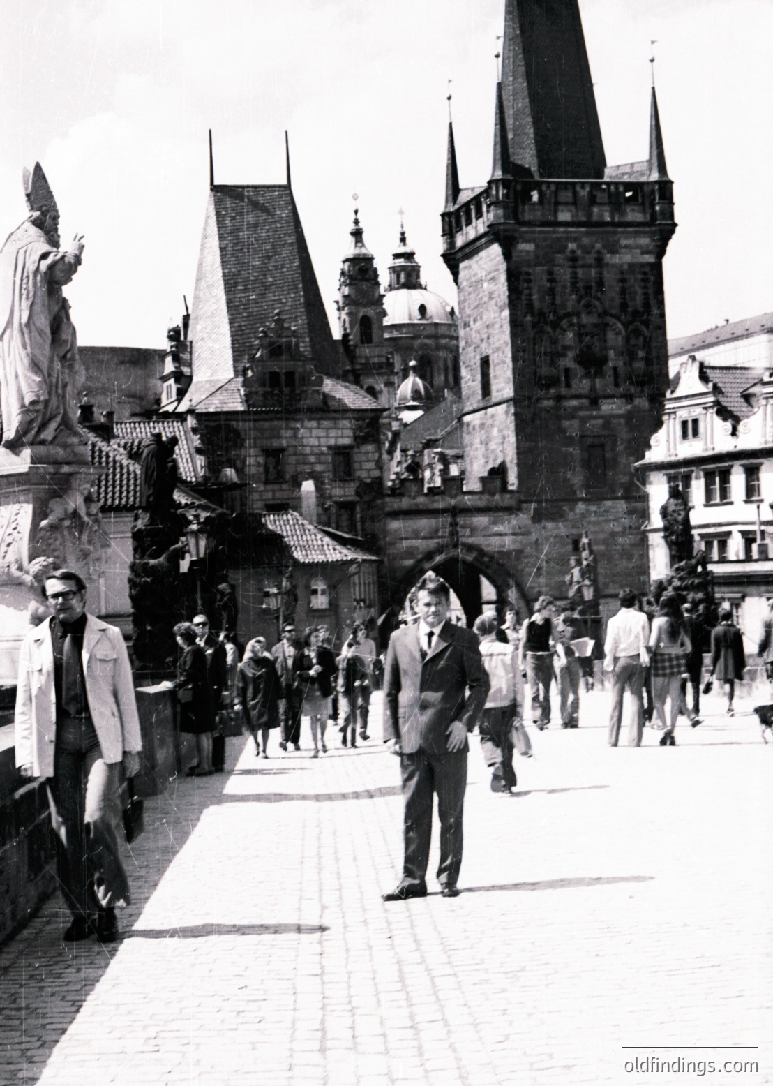 Black-and-white street scene at Prague’s Charles Bridge () during the 1960s (). Gothic towers, statues, and historic stone arches frame pedestrians in classic European attire. Crowds gather near the Old Town () under clear skies.
